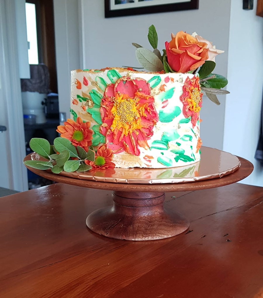 A colorful flower-adorned cake on a wooden cake stand, decorated with large orange and pink flowers and green leaves, set indoors on a wooden table.