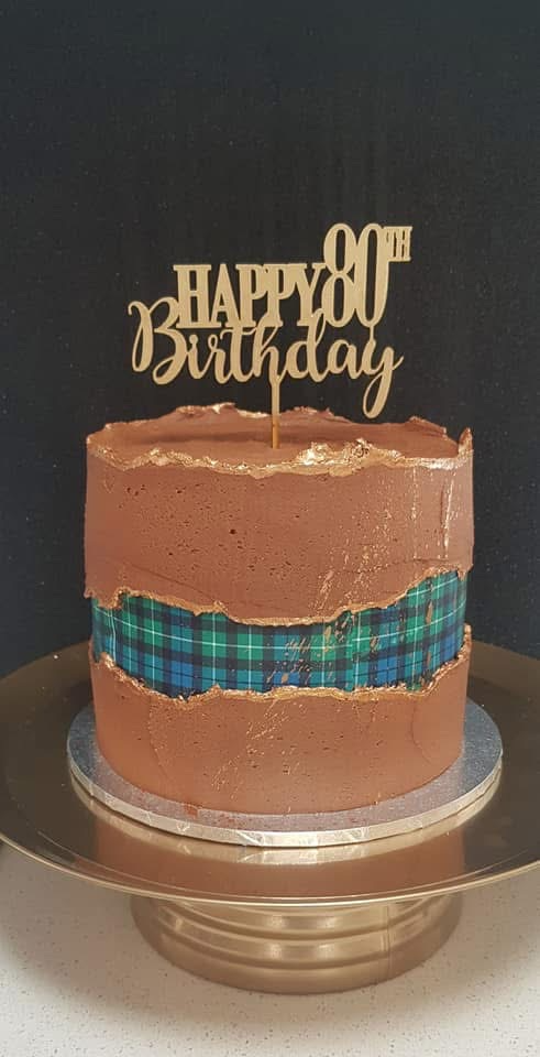 A chocolate birthday cake with a blue and green tartan band around the middle and a gold 'Happy 90th Birthday' topper on top, placed on a silver cake stand.