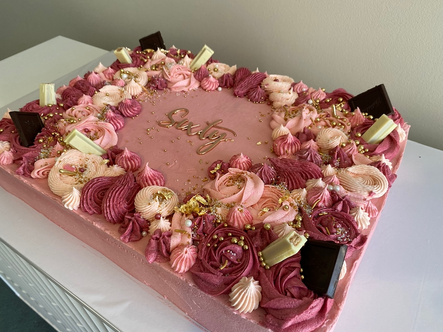 A heart-shaped pink birthday cake decorated with pink and white whipped cream, chocolates, gold beads, and the name "Euly" written in gold lettering.