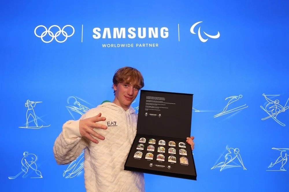A young athlete in a white EA7 hoodie holding an open collector's box of Samsung Olympic commemorative pins against a blue backdrop featuring Samsung and Olympic logos.