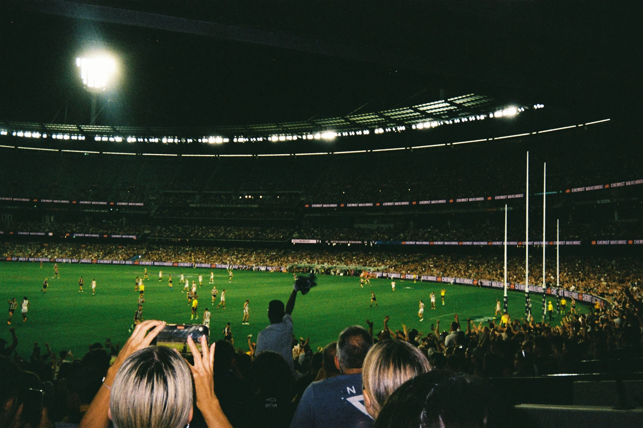 Nighttime AFL game in a stadium with bright lights, players on the field, and spectators in the stands taking photos and cheering.