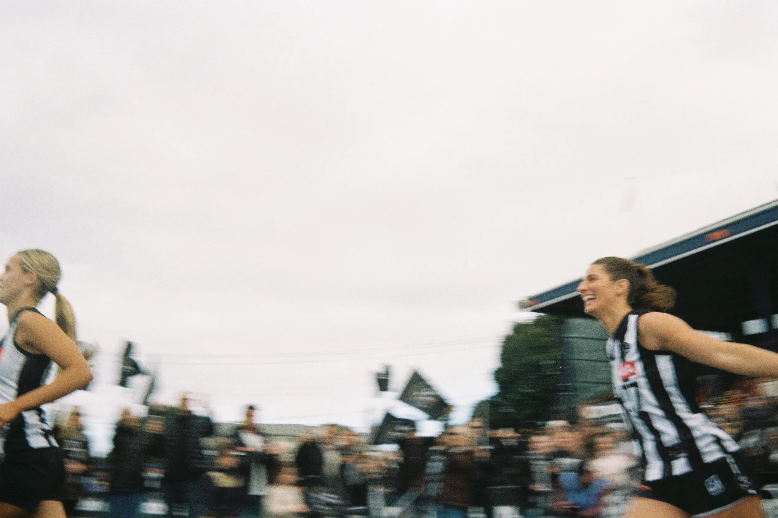 Two women wearing black and white striped sports uniforms running in a race, smiling, with a crowd and trees in the background.