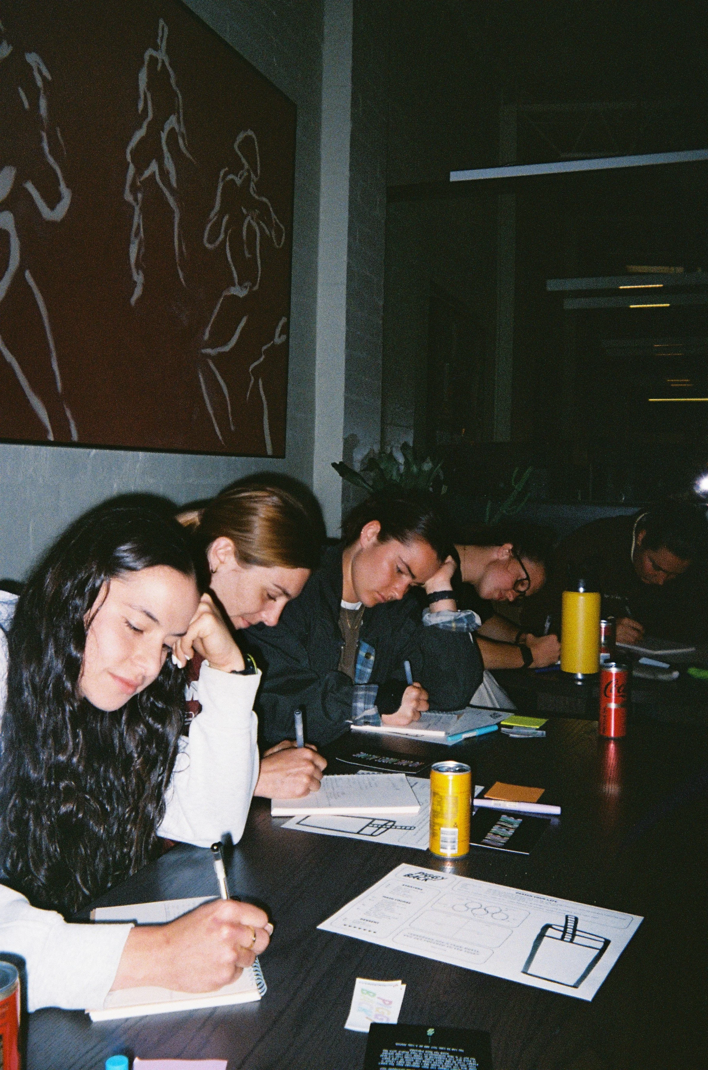 Group of five people sitting at a table, taking notes with pens, in a dimly lit room.
