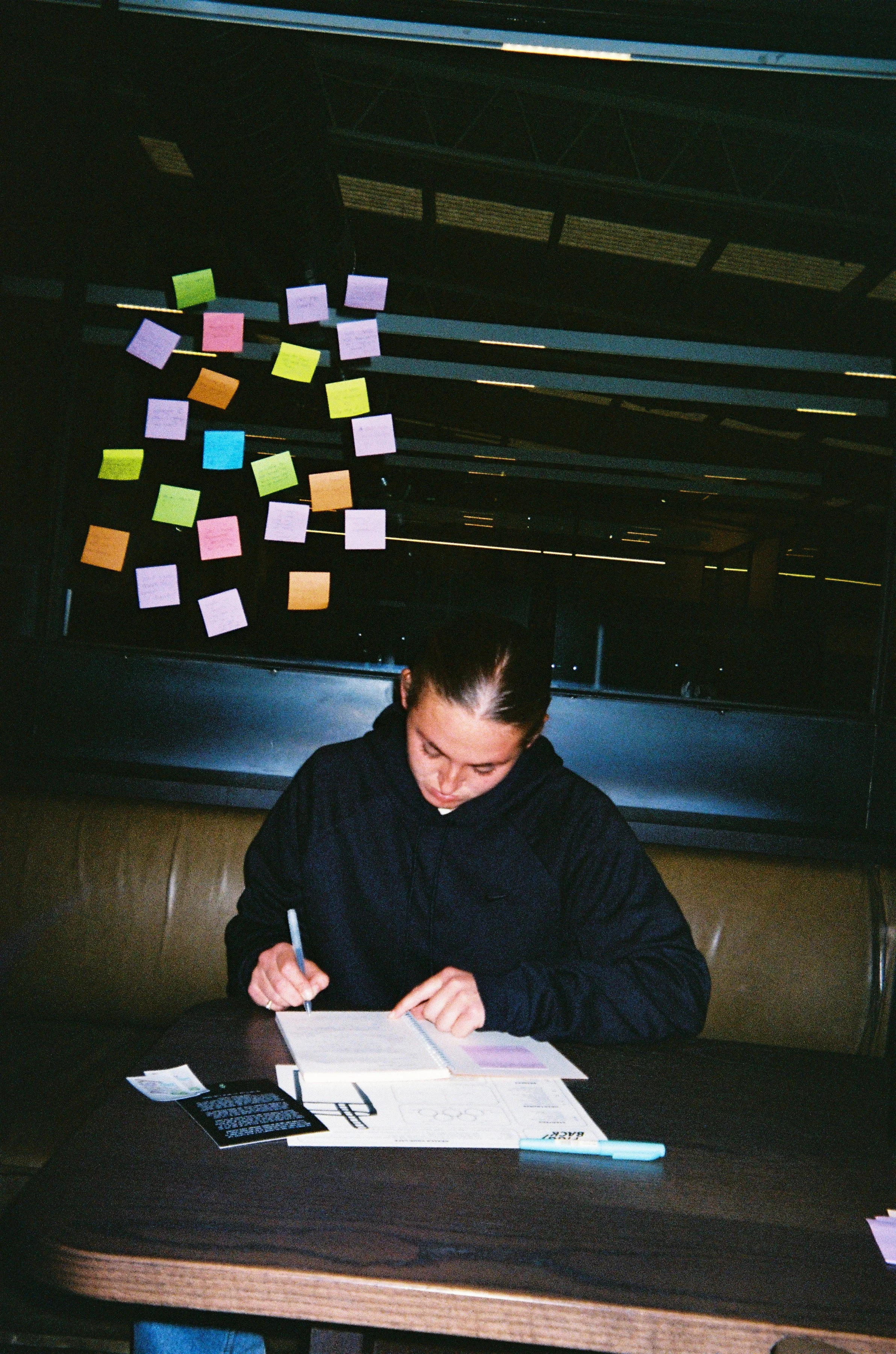 A woman sitting at a table, writing in a notebook with colorful sticky notes on the window behind her.