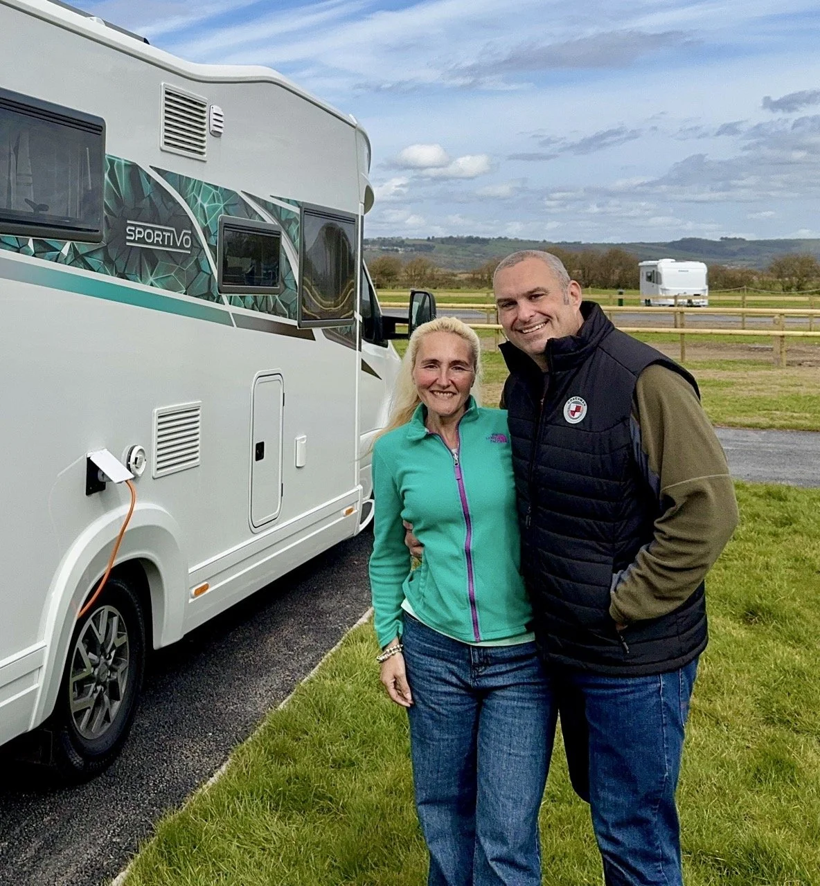 A smiling woman and man standing outdoors near a white RV with a scenic landscape in the background.