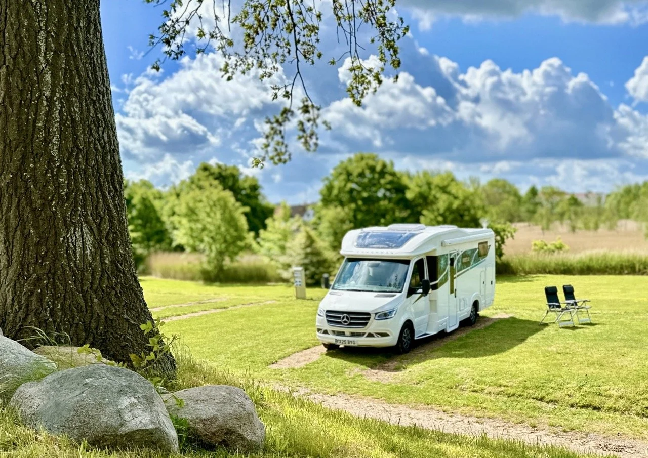 A white camper van parked on a grassy area under a large tree with chairs nearby, with a blue sky and fluffy clouds in the background.