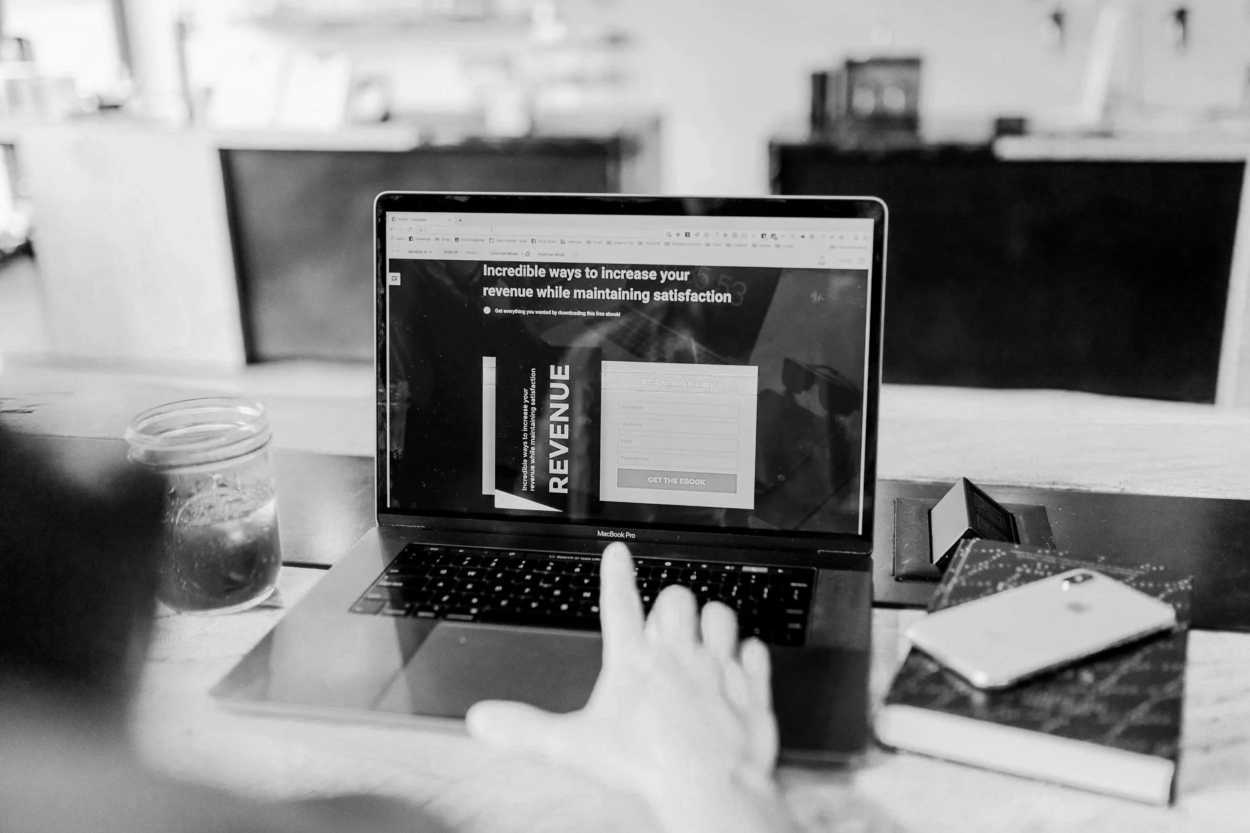Black and white photo of a person using a MacBook Pro to view a website about increasing revenue, with a glass jar of water, a smartphone, a notebook, and a small stand on a wooden table.