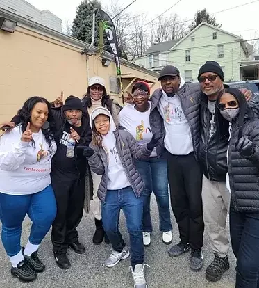 Group of seven people standing outside, smiling and posing for a photo, some wearing jackets and hats, in a residential neighborhood with houses in the background.