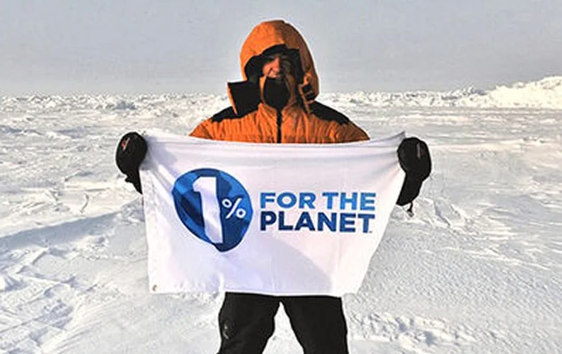 Person wearing an orange jacket and black gloves holding a banner in a snowy landscape that reads '1% for the Planet'.