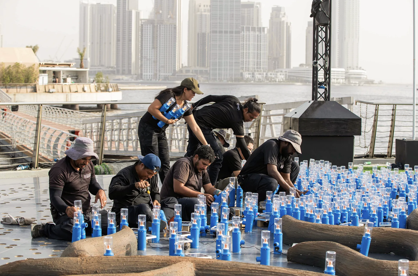 A group of five people working together outdoors, with some holding a wooden plank, in a rustic woodworking area with trees and a shed in the background.