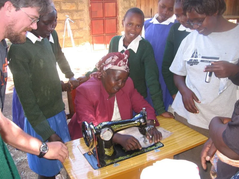 An elderly woman sewing on a vintage black machine while young students and a teacher observe around her in a classroom.