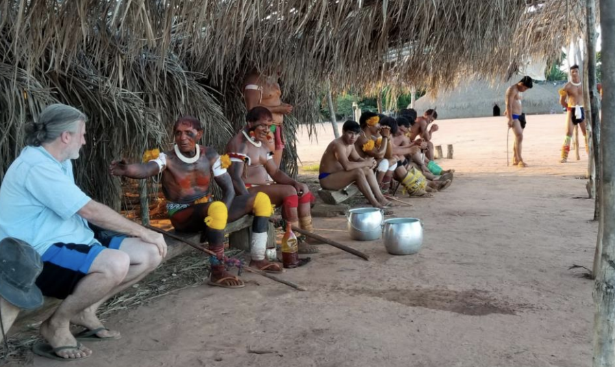 A group of indigenous people sitting under a thatched roof, with some holding cups or bowls, and a foreign man sitting nearby, possibly observing or participating.