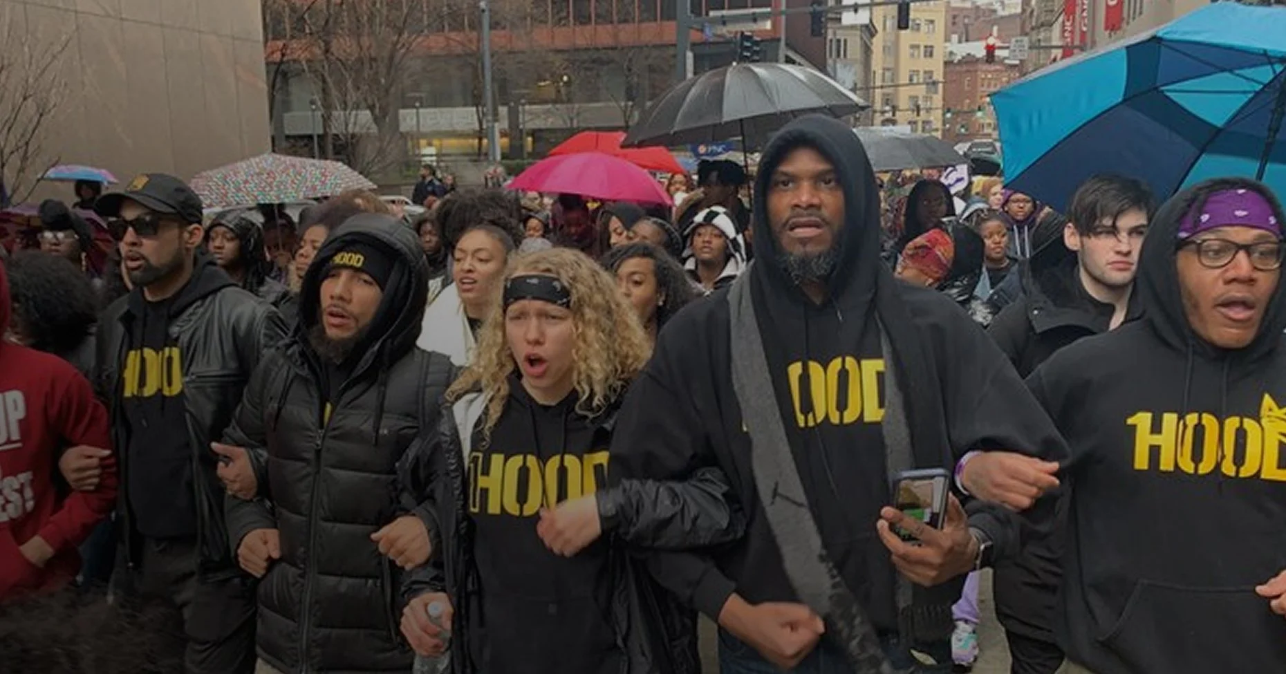 A group of diverse people, including men and women, participating in a protest or march. Many are wearing black hoodies with yellow text that says 'Hood'. Some are holding umbrellas due to rain, and some appear to be singing or chanting.