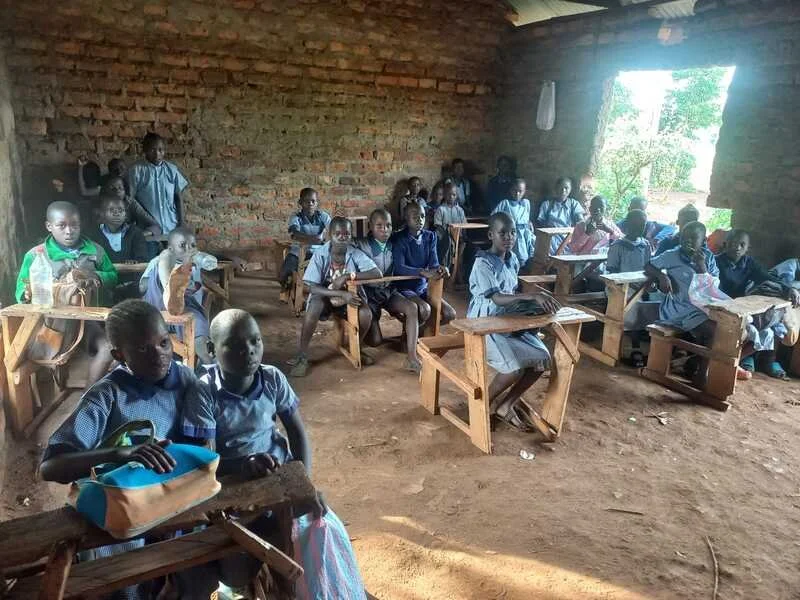 Group of children in school uniforms sitting at wooden desks inside a classroom with brick walls, some students have backpacks, sunlight coming through an open window.