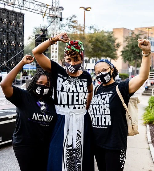 Three women wearing black 'Black Voters Matter' T-shirts and face masks, raising their fists in a fist pump gesture outdoors during daytime.