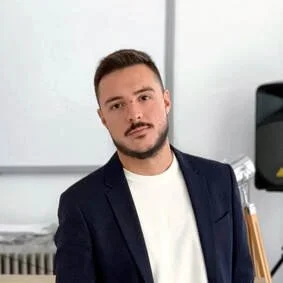 Portrait of a young man with short brown hair and a beard, wearing a dark blazer over a white shirt, standing in a modern office setting.