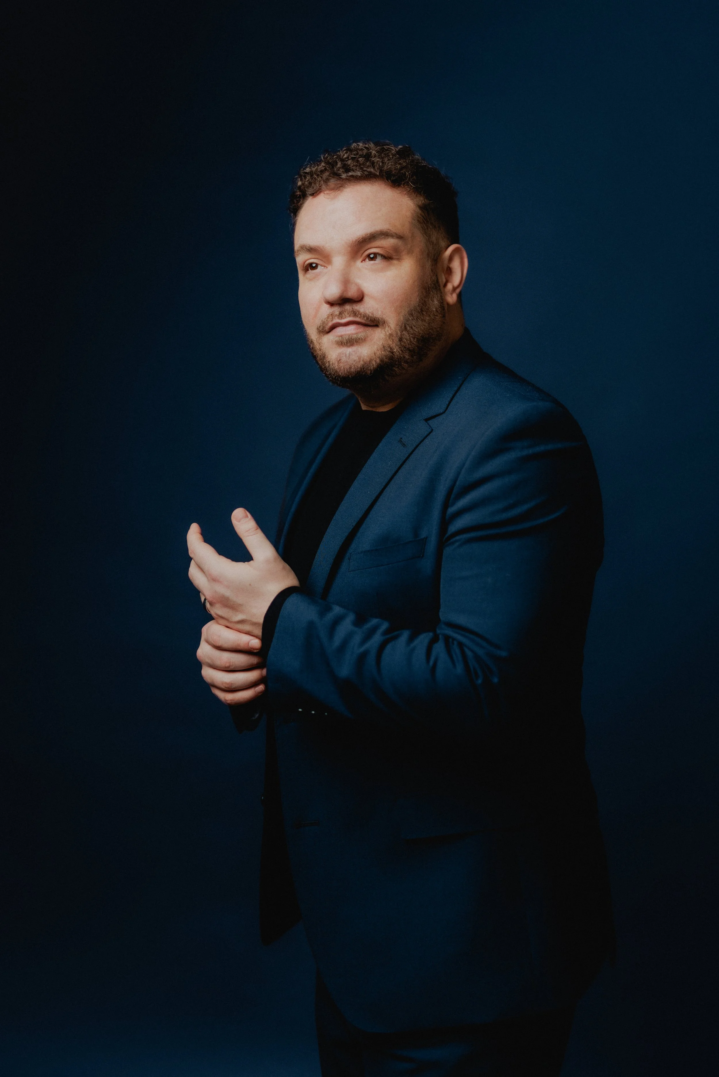 Portrait of a man in a dark blue suit with a black shirt, standing against a dark blue background, looking off to the side with a slight smile.
