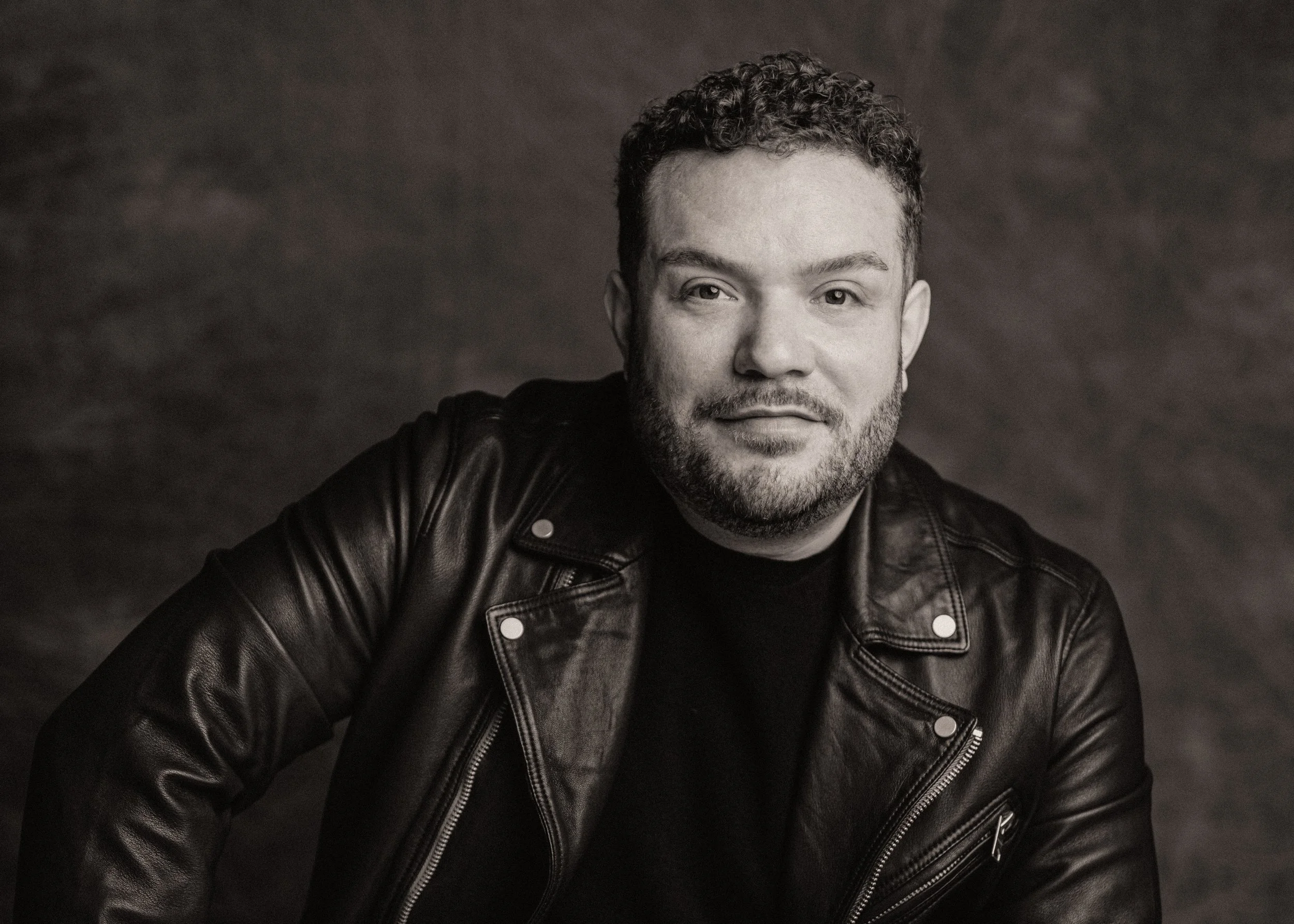 Black and white portrait of a man with curly hair and a beard, wearing a leather jacket and smiling slightly, posed against a dark textured background.