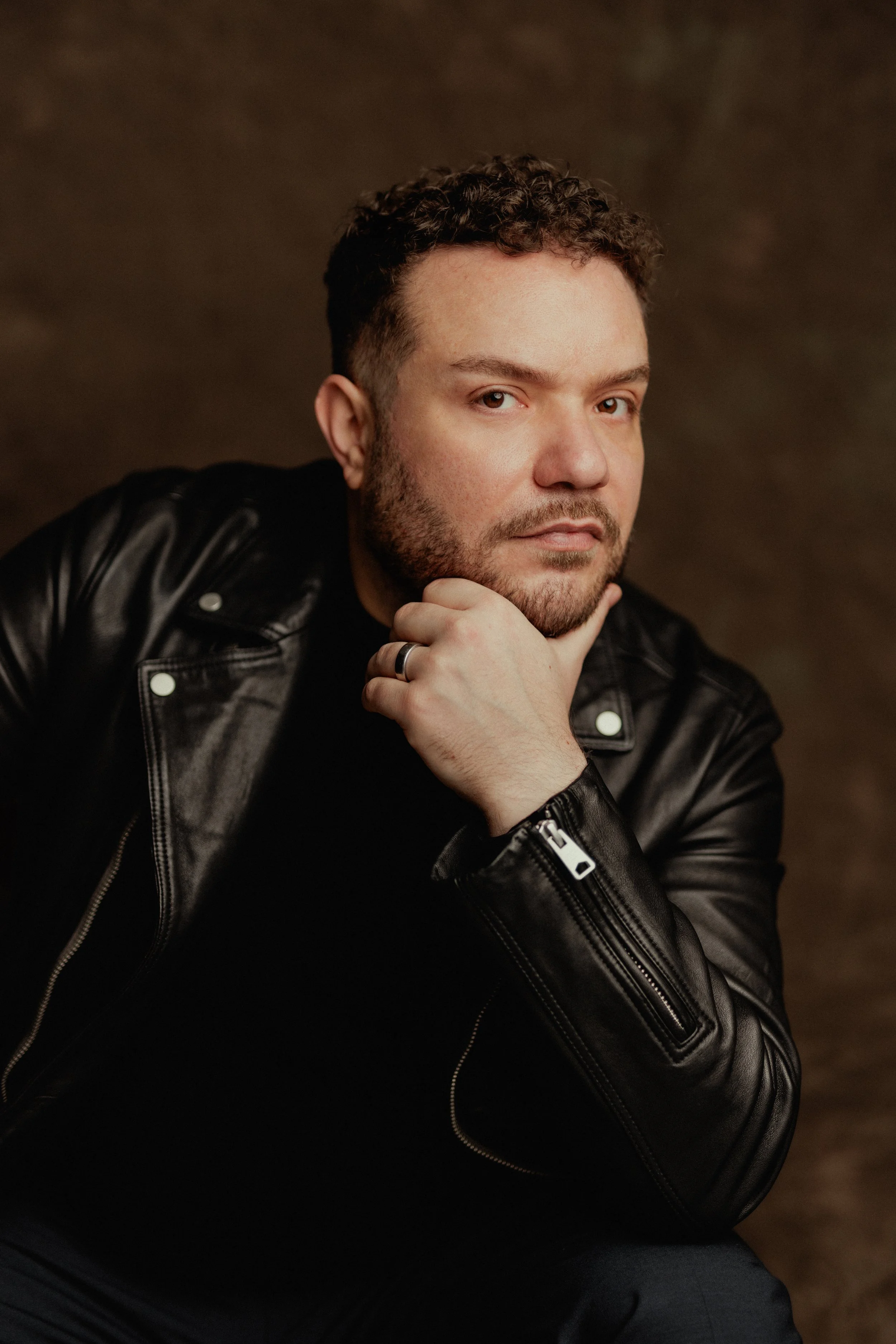 A man with curly hair and a beard wearing a black leather jacket, sitting with his hand on his chin, looking at the camera.