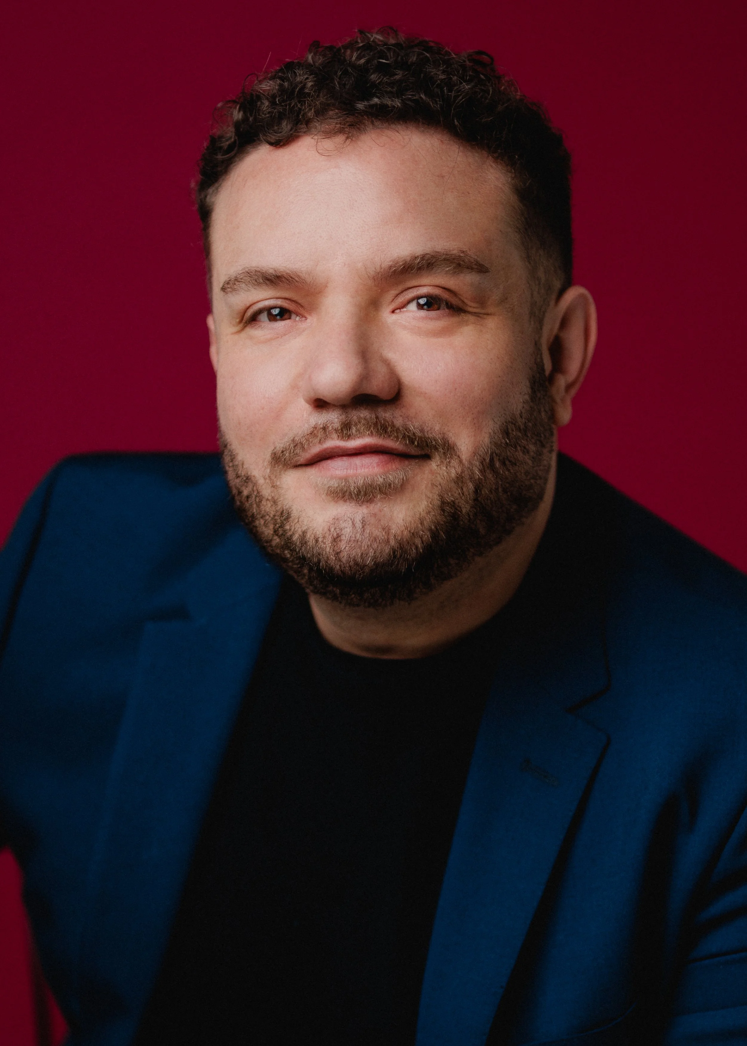 A man with short curly hair and a beard, wearing a dark shirt and a blue blazer, smiling slightly, against a red background.