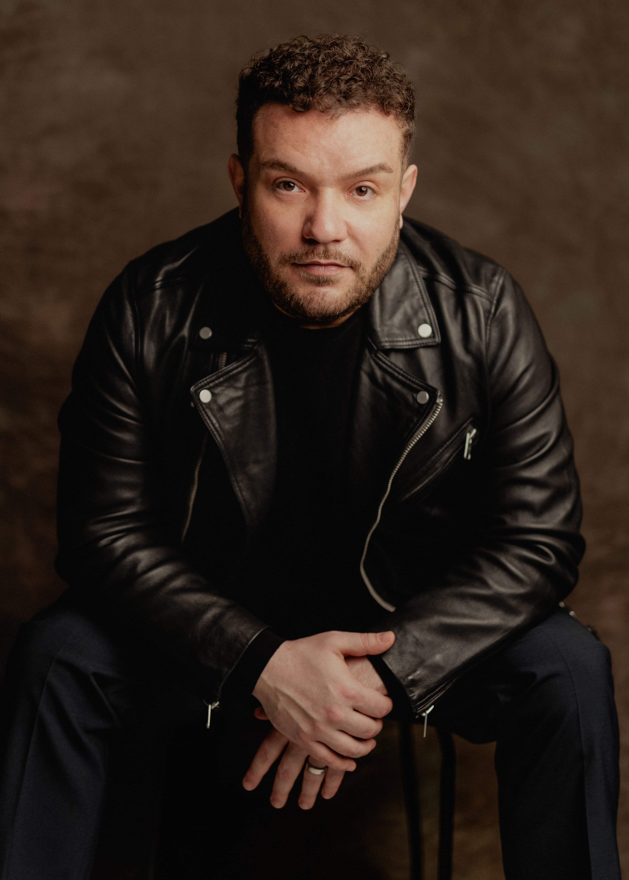 A man with curly hair and beard wearing a black leather jacket and black pants, sitting against a brown background, looking at the camera.