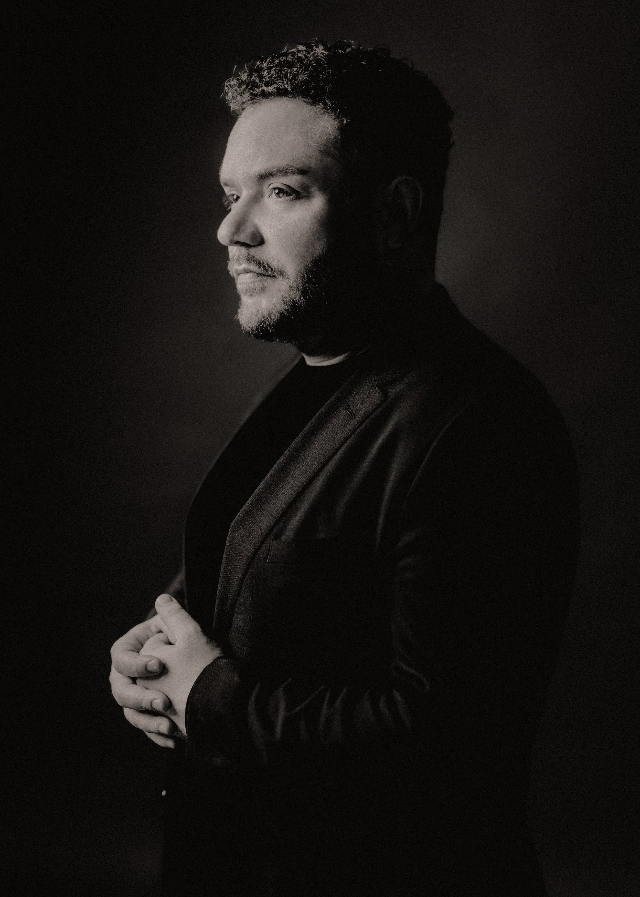 Black and white portrait of a man with short, curly hair and a beard, wearing a dark suit jacket and shirt, with hands clasped in front of him, looking to the left against a dark background.