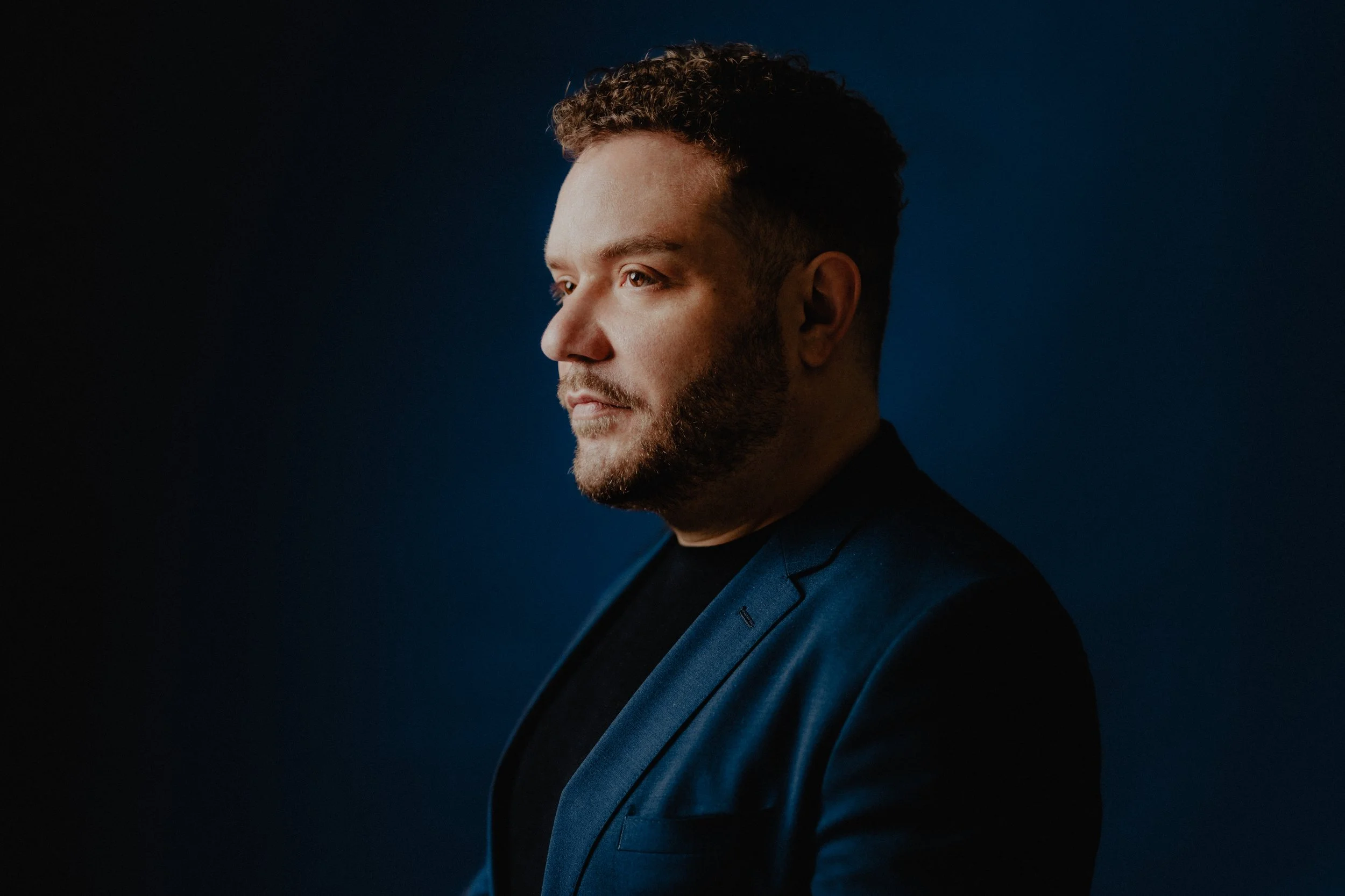 A young man with curly hair and a beard in profile view, wearing a dark blue blazer, against a dark blue background.
