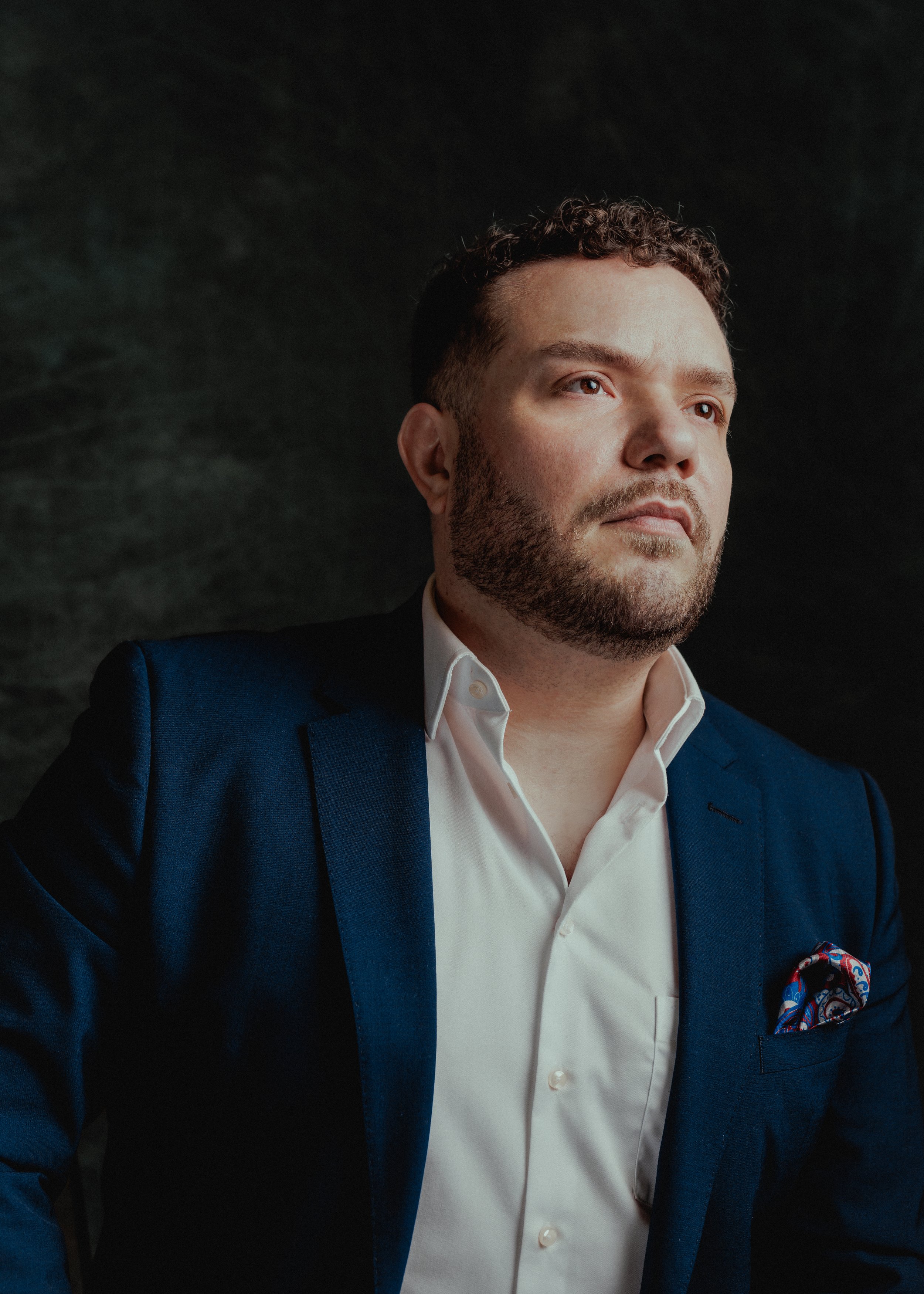 A man with a beard and curly hair, wearing a blue suit and white shirt, looking thoughtfully to the right, against a dark background.