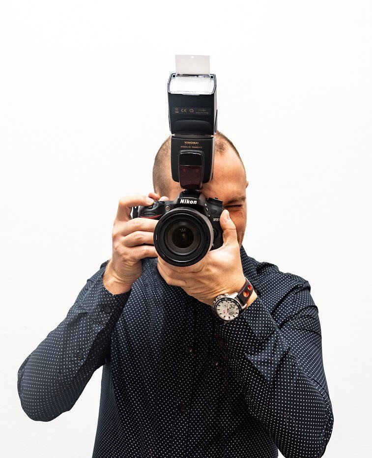 A man taking a photograph with a camera, with a flash attached on top, against a plain white background.