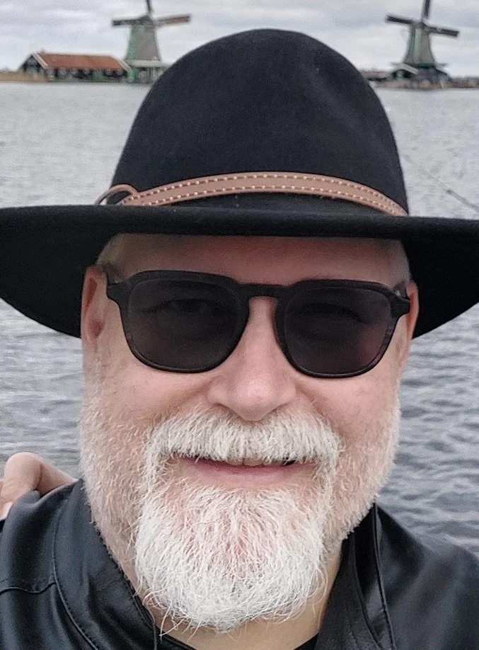 A smiling man with a white beard and mustache wearing sunglasses, a black hat with a leather band, and a black leather jacket stands outdoors near water with windmill structures in the background.