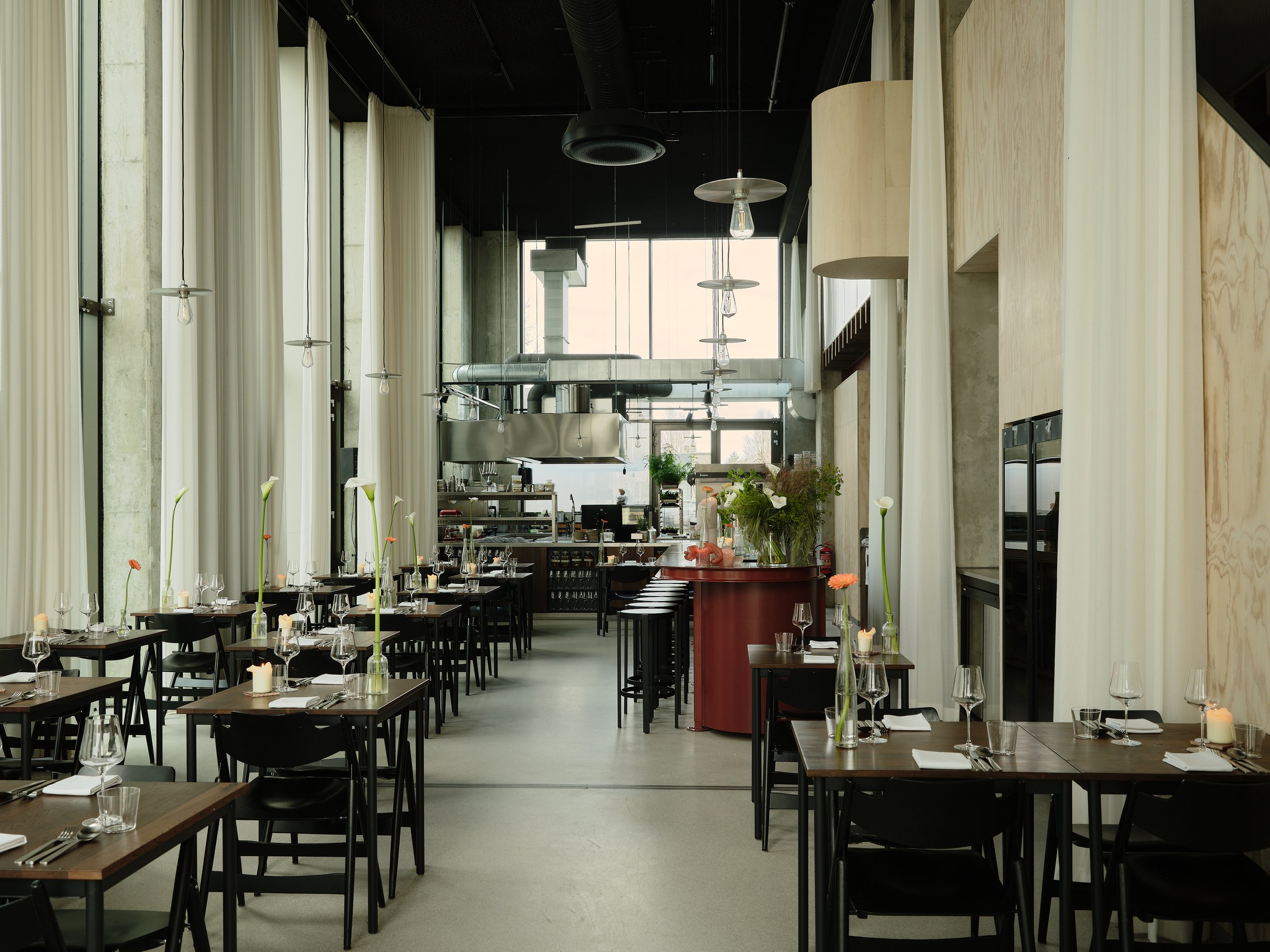 Interior of a modern restaurant with black tables, chairs, tall windows with white curtains, and minimalist decor. Centered is a red bar table with floral arrangements, and the background features an open kitchen area with industrial stainless steel equipment.