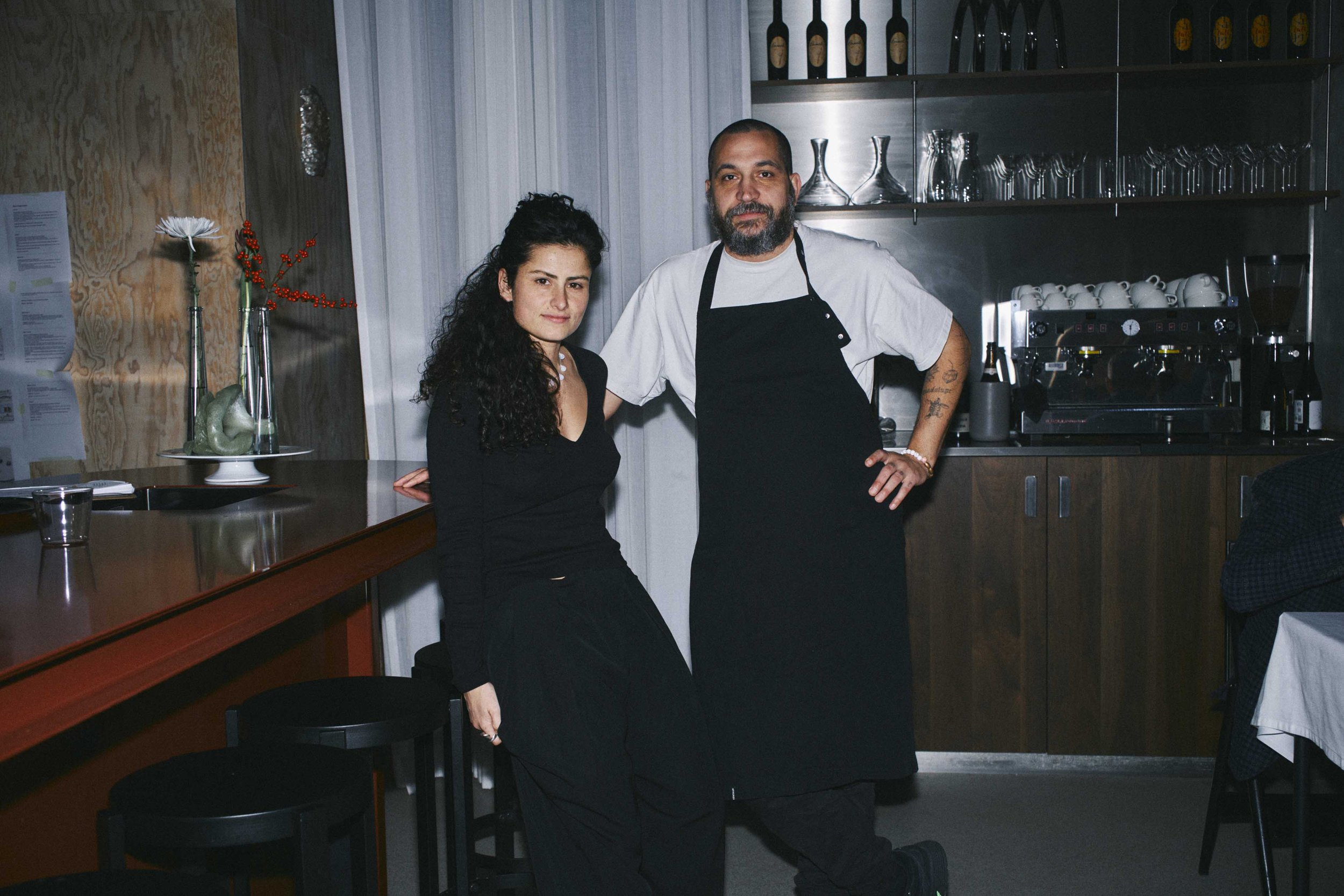 Frederieke Bloem and Ivan Beusink, owner and chef of Copain. The woman with long curly hair dressed in black and the man with a beard wearing a white T-shirt and black apron, standing in front of a kitchen area with glasses and coffee equipment.