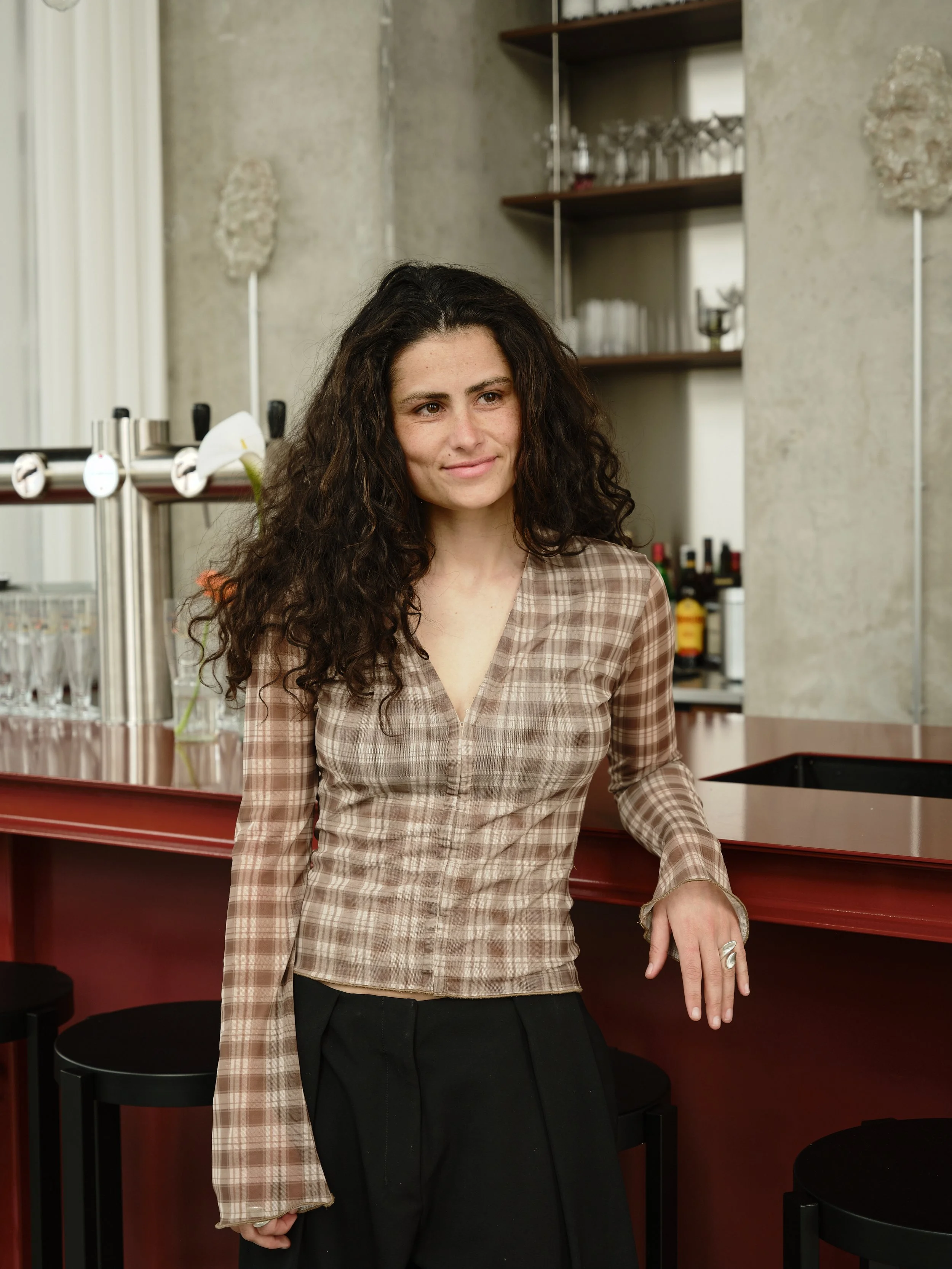 A woman with long, curly dark hair leaning against a bar counter in a modern interior space.