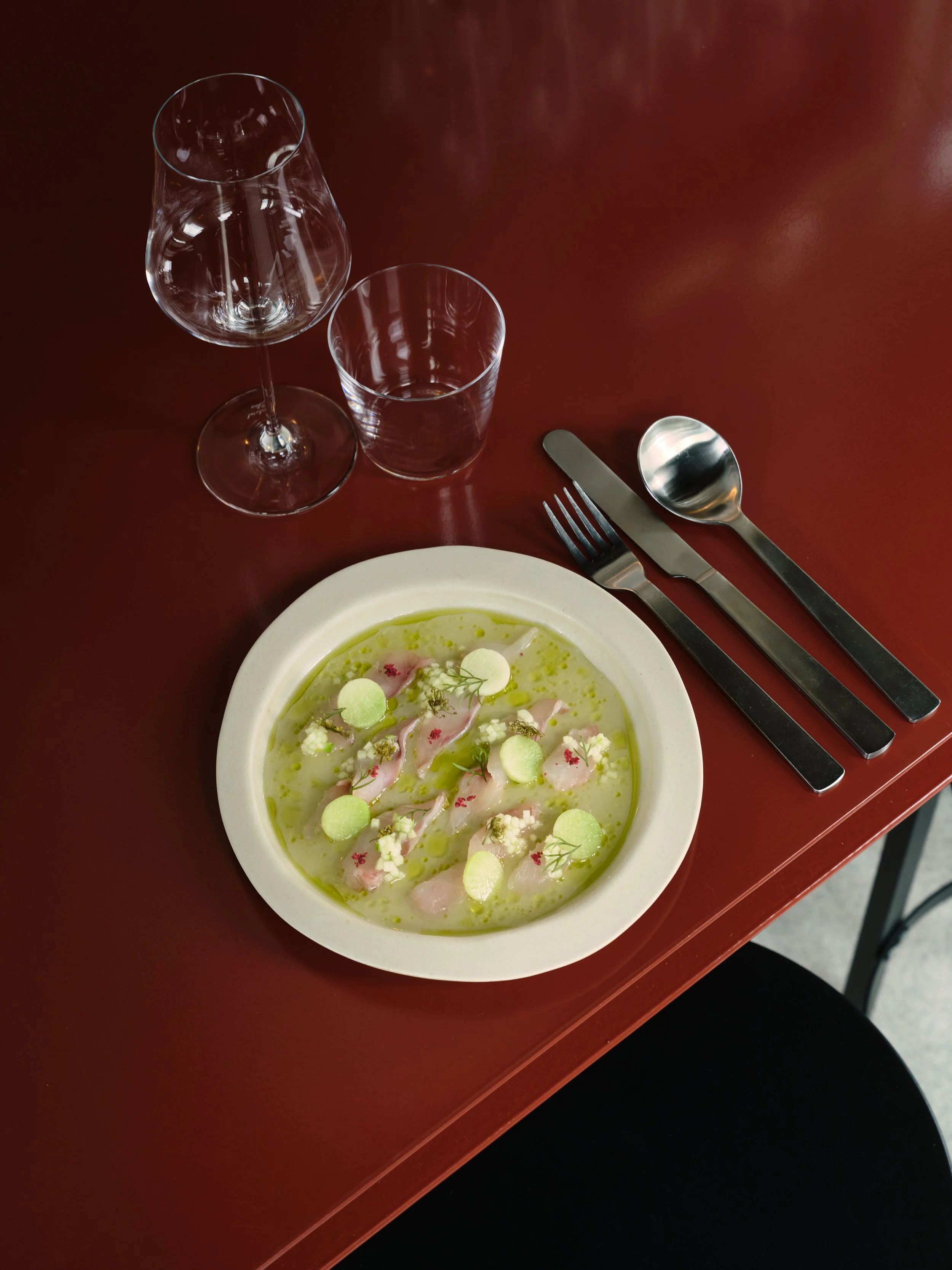 A white plate with a fine dining dish containing slices of fish, garnished with herbs and small green and white spheres, served on a red table next to a fork, knife, spoon, water glass, and wine glass.