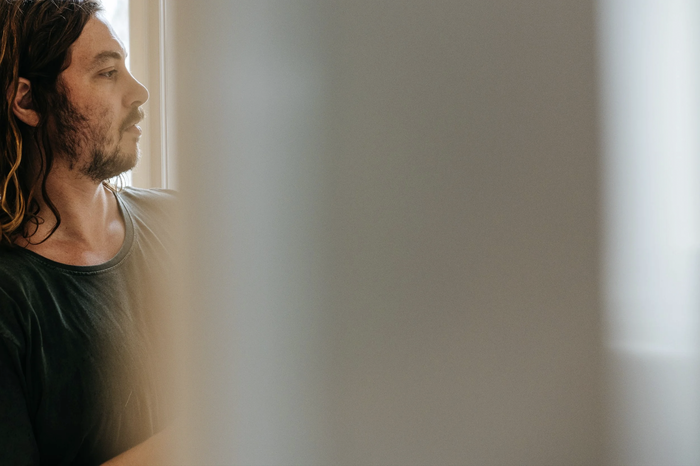 A man with long hair and a beard looking out a window.