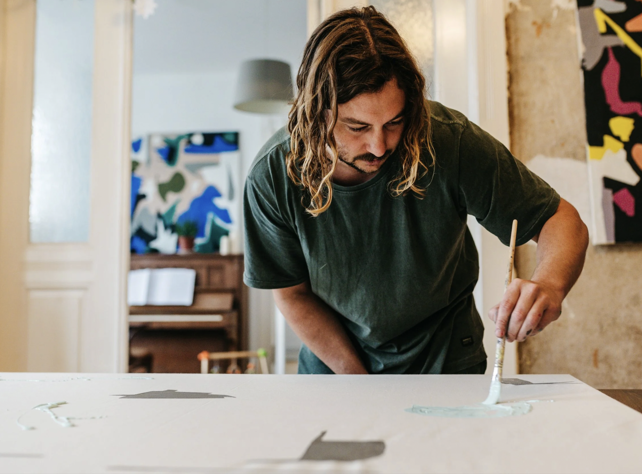 Man with long, wavy hair wearing a dark t-shirt, painting on a large canvas with a brush in an art studio.