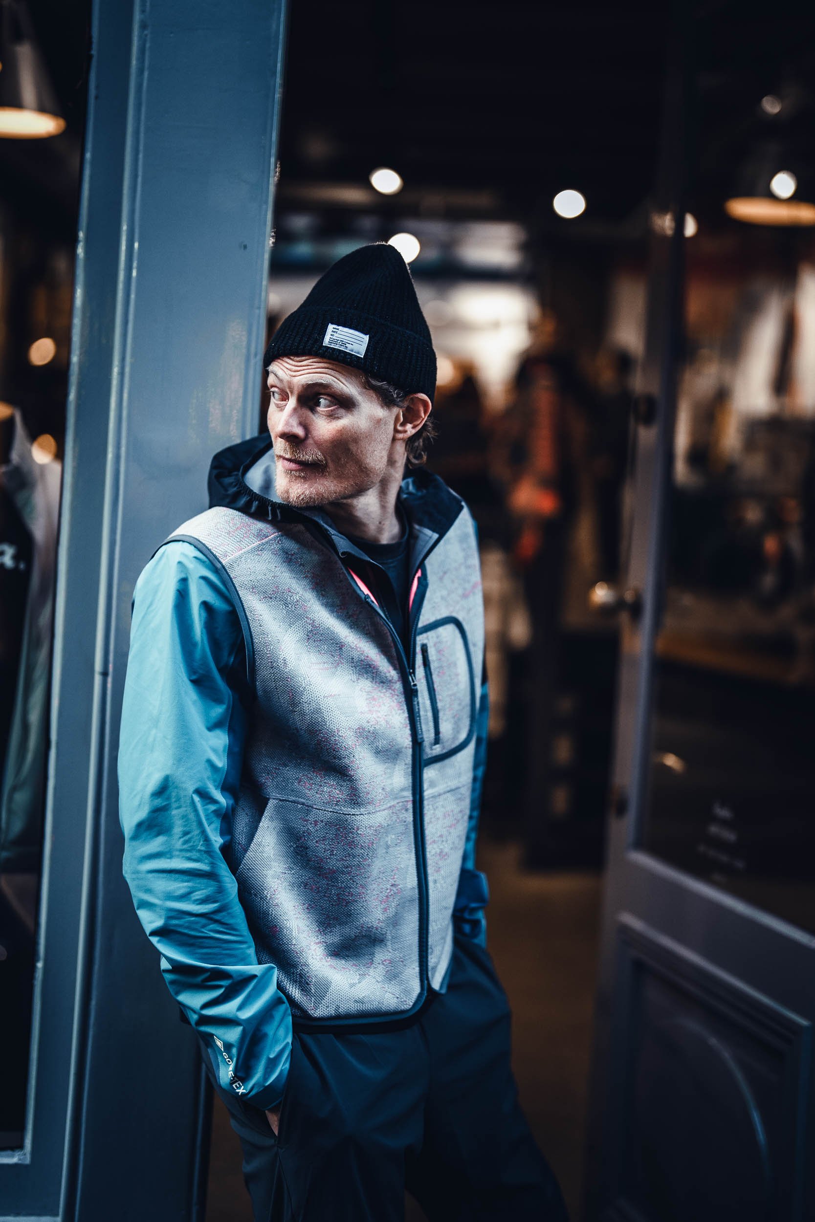 A man wearing a black beanie, a grey and blue jacket, posing indoors in a dimly lit space, looking to his left.