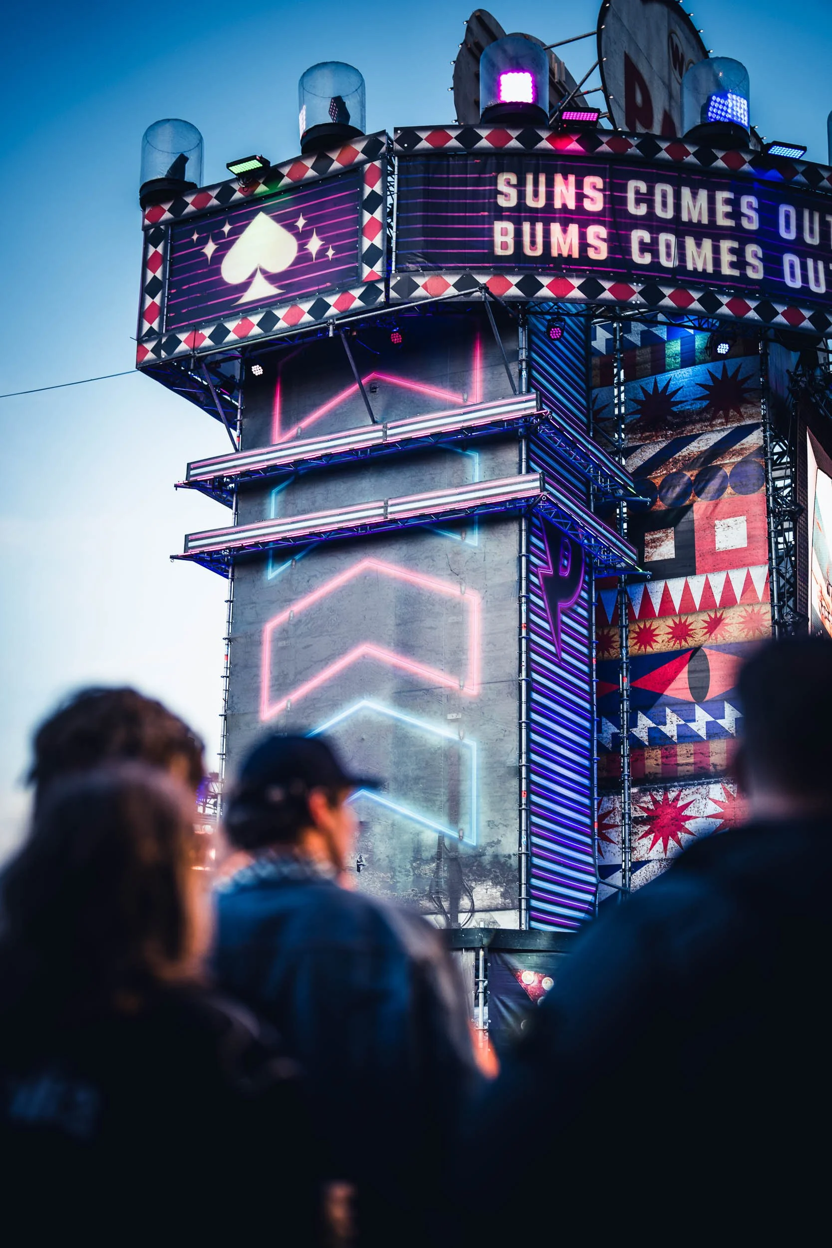 Nighttime scene at a carnival with a neon-lit amusement ride or game structure featuring bright pink, blue, and purple lights, and a sign that reads "Suns comes out, Bums comes out." A group of people is in the foreground observing the attraction. 