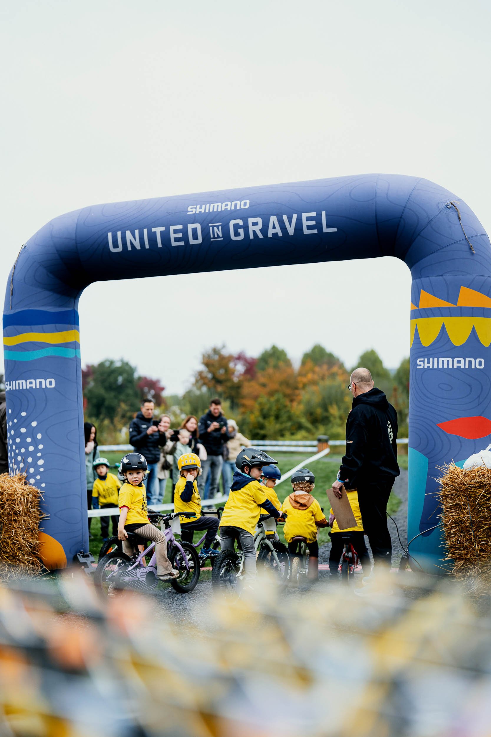 Children in yellow shirts on bicycles at the starting line under a large blue archway labeled 'United in Gravel' with spectators in the background.
