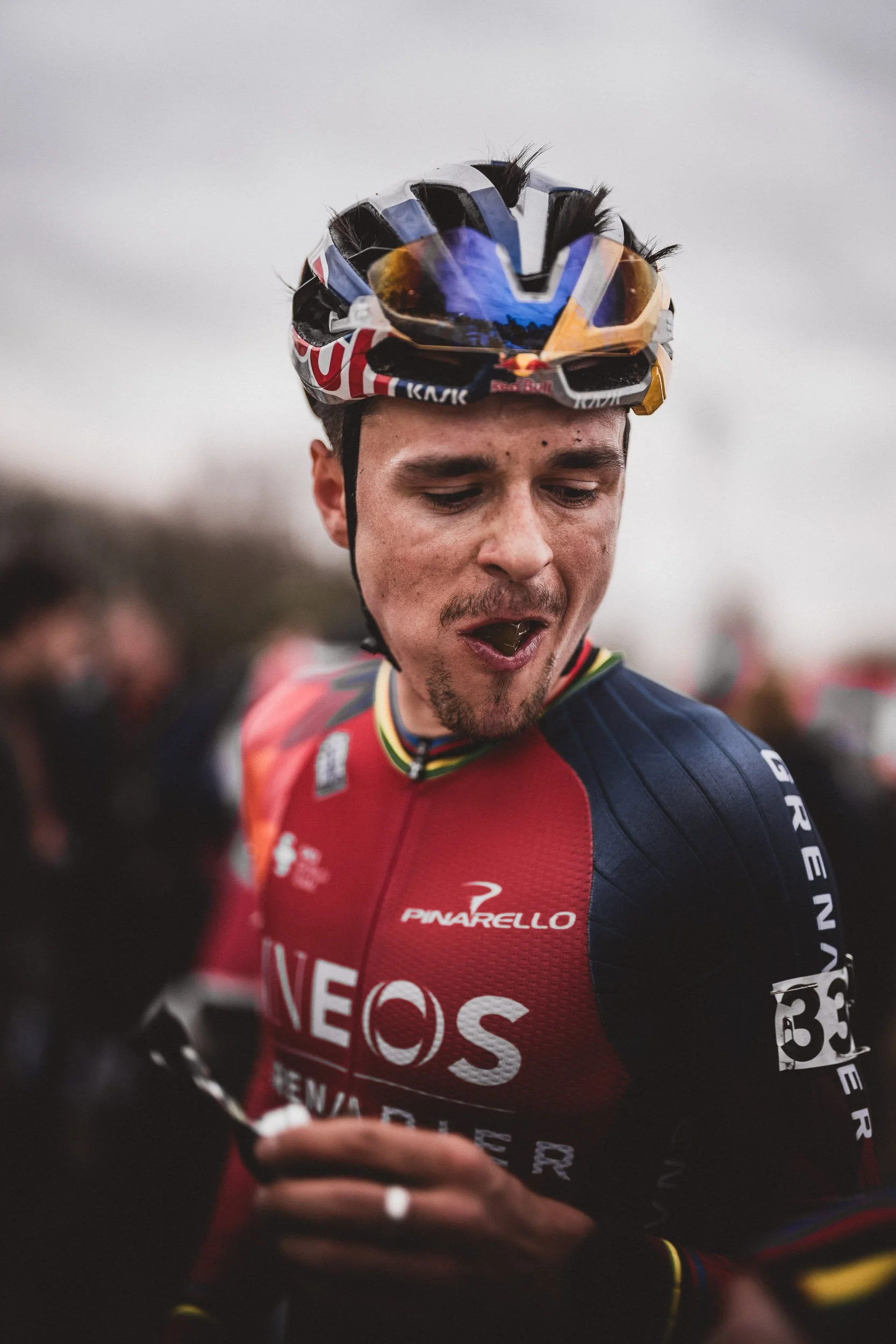 A male cyclist wearing a red and navy cycling jersey with logos, a colorful helmet with a visor, and sunglasses, appears to be celebrating or reacting during a race. (Tom Pidcock) (Ineso Grenadiers)