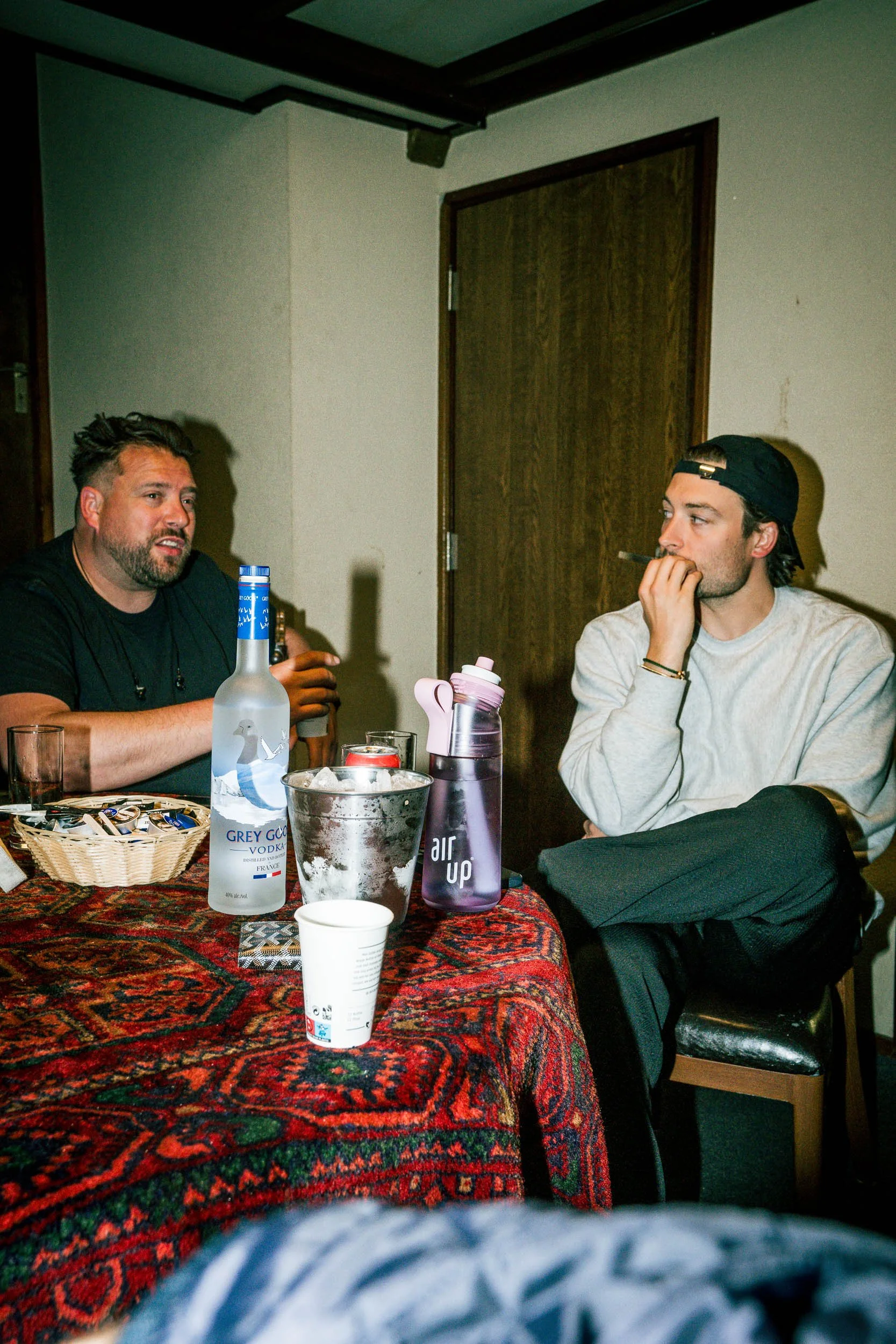Two men sitting at a table having a conversation indoors. One man has a beard and is wearing a black shirt, the other is wearing a light grey sweatshirt and a backwards cap.   (Viktor Verhulst)