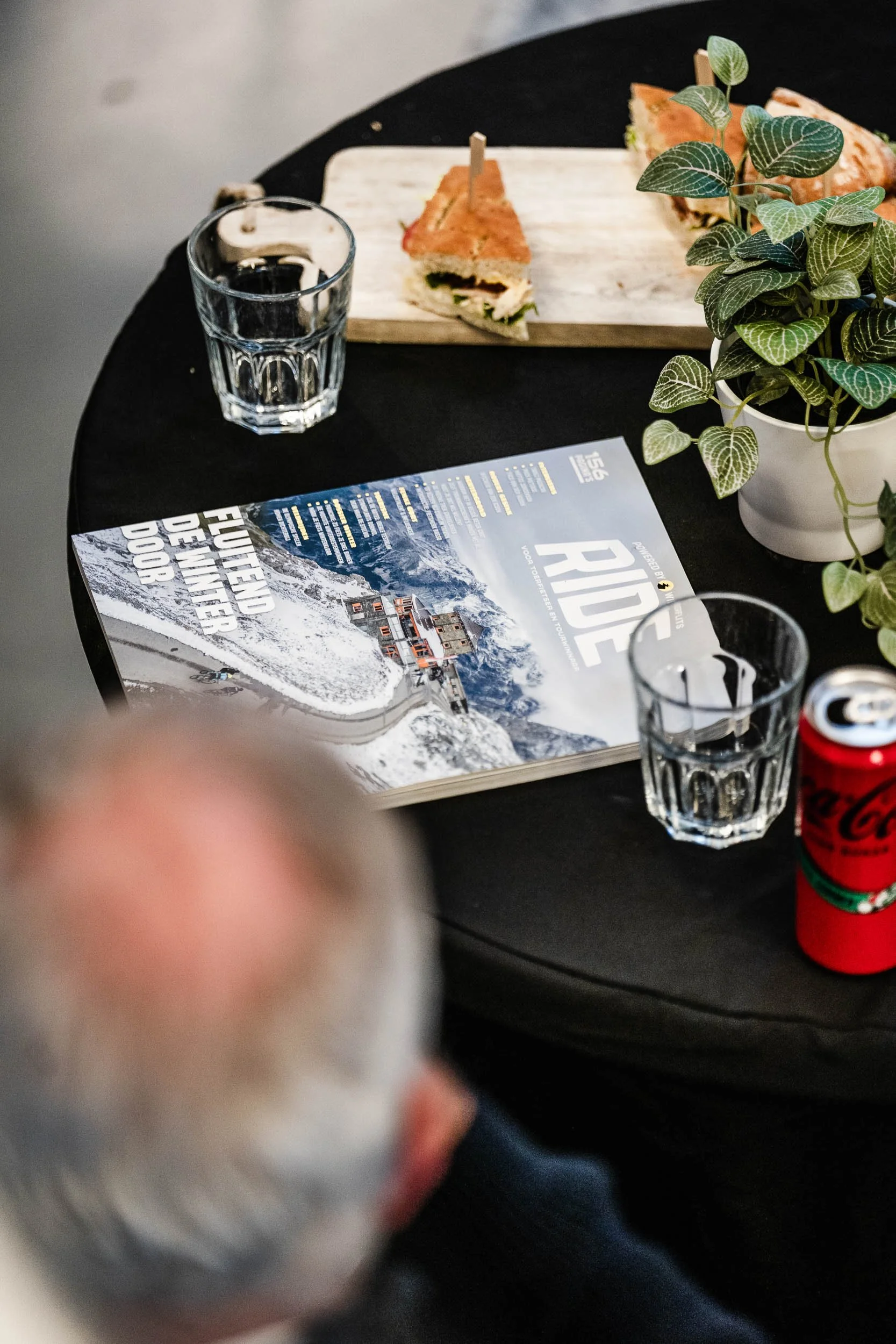 A table with a mountain-themed magazine, two glasses of water, a plant in a white pot, a can of Coca-Cola, and a wooden cutting board with sandwiches. A person with gray hair is blurred in the foreground.