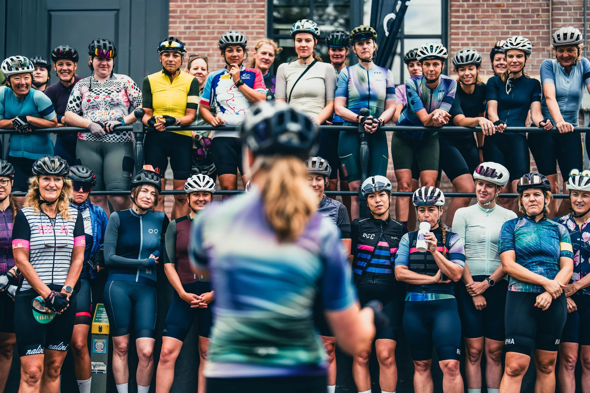 A group of women in cycling gear gathered outdoors, wearing helmets, listening to a woman speaking in front of them.