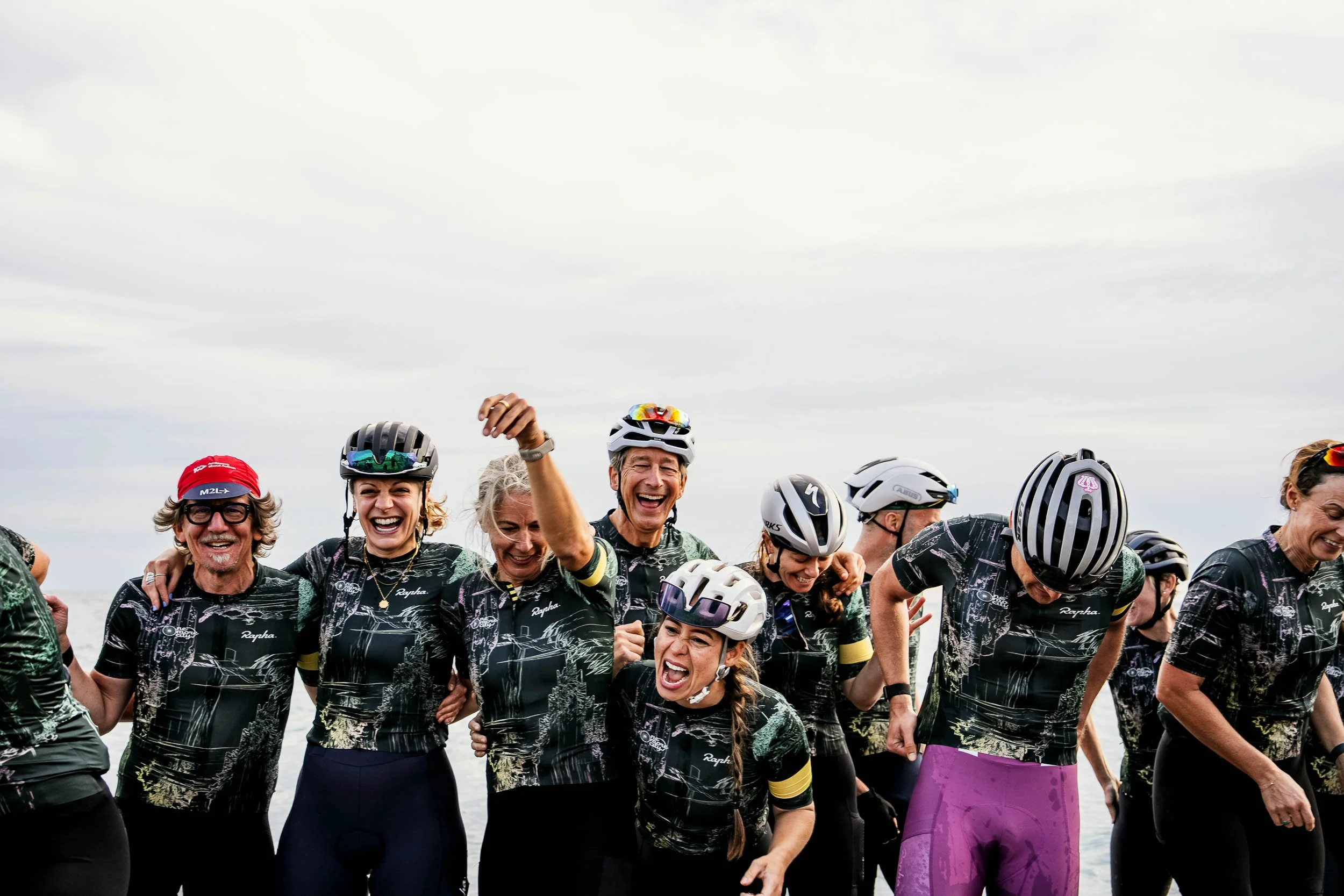 A group of cyclists wearing matching black jerseys with a mountain and river design, celebrating together, some with helmets, smiling and laughing. (One More City  Rapha.cc)