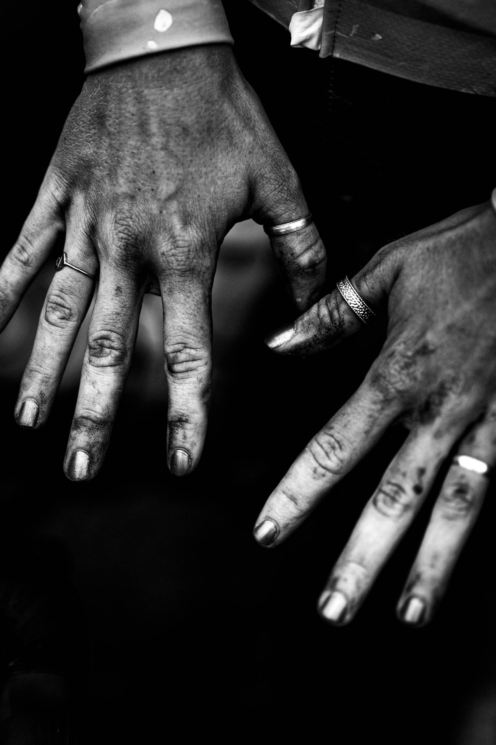 Black and white close-up of two hands with rings, displaying dirty fingernails and textured skin, with one hand positioned above the other.