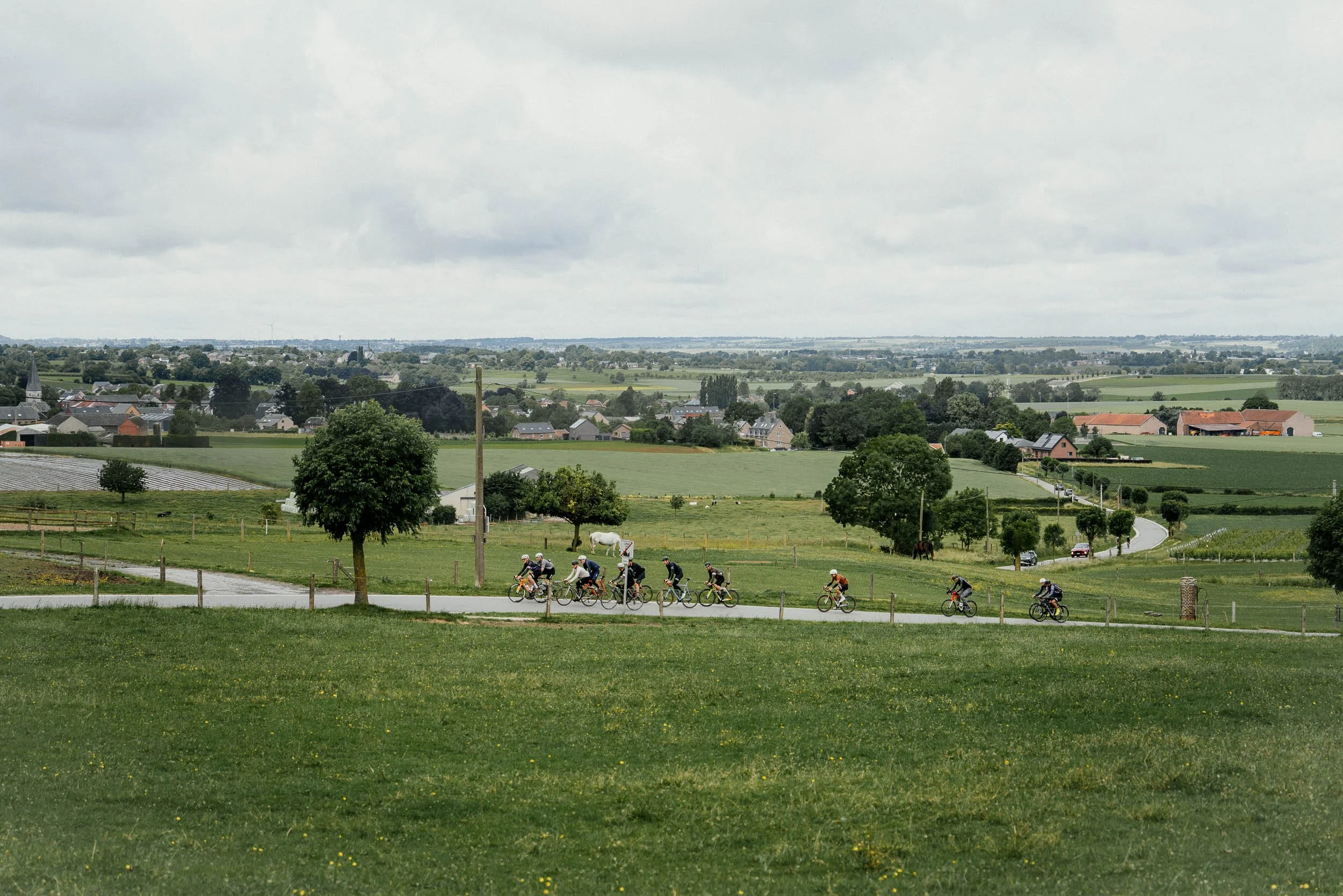 Group of cyclists riding on a rural road through a green countryside with trees, fields, and a cloudy sky