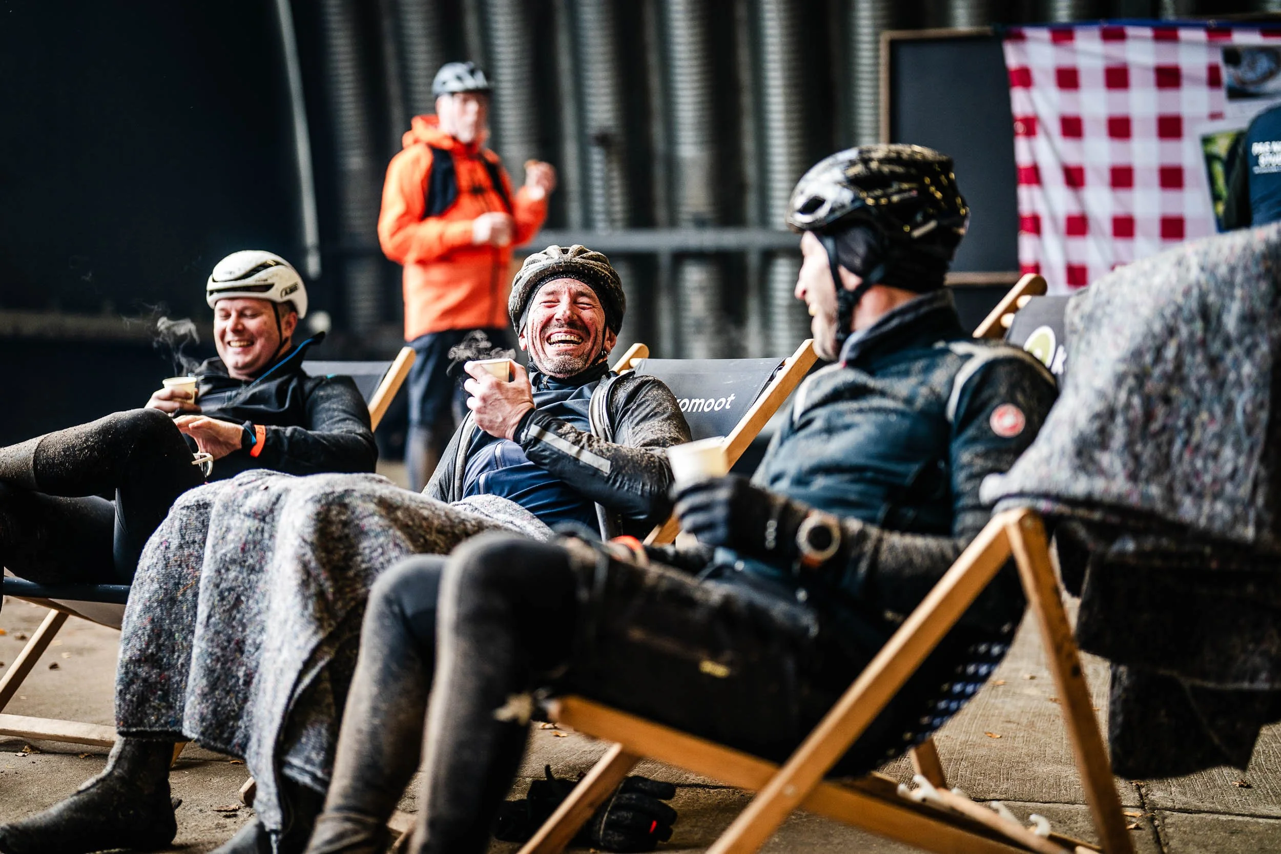 Four men in cycling gear and helmets relaxing on lounge chairs, laughing and enjoying drinks, with one man showing a cheerful expression, in an indoor setting with industrial elements and a person in the background.