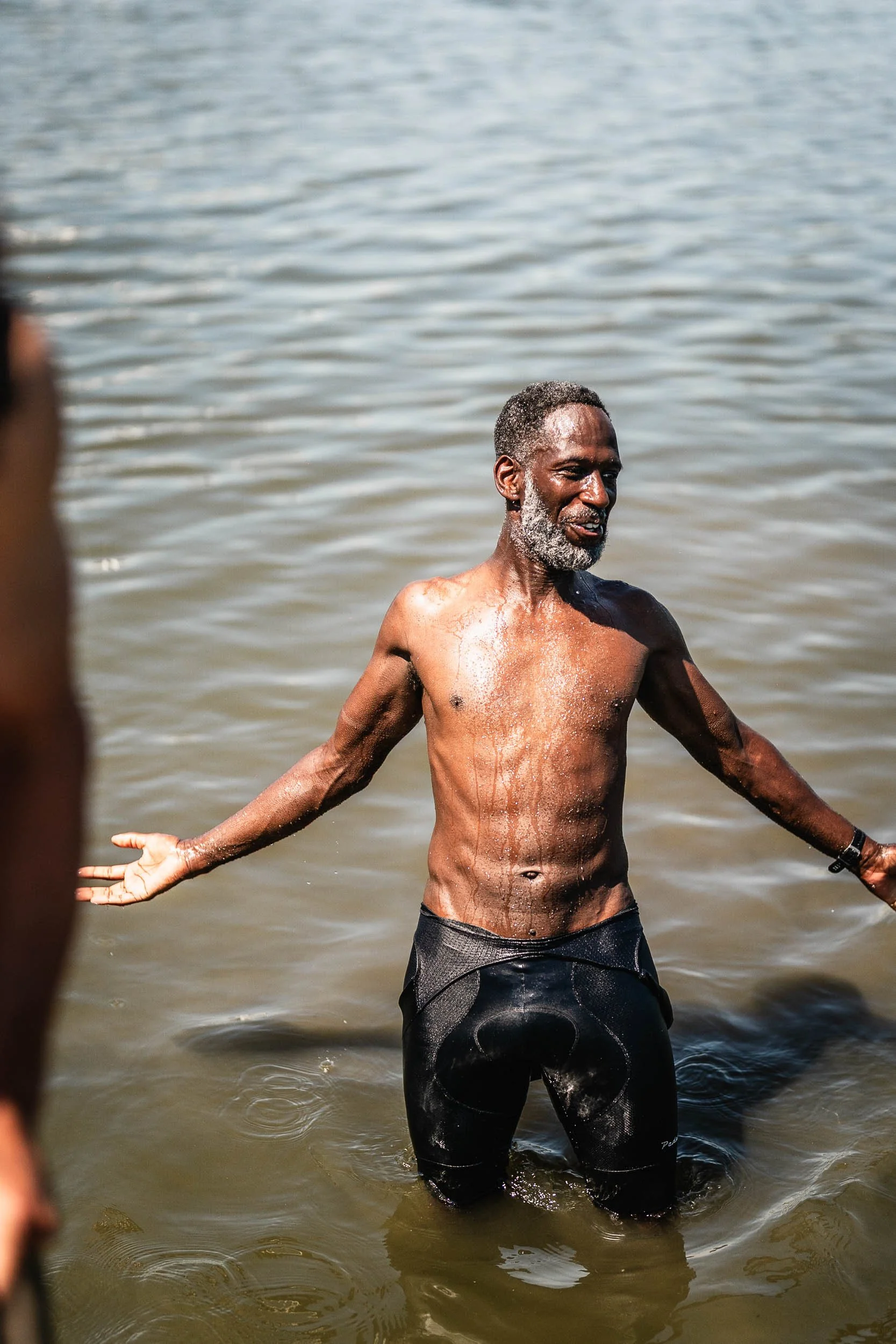 A shirtless older man with a beard standing waist-deep in water, smiling and gesturing with arms outstretched. (Patta.nl Rapha)