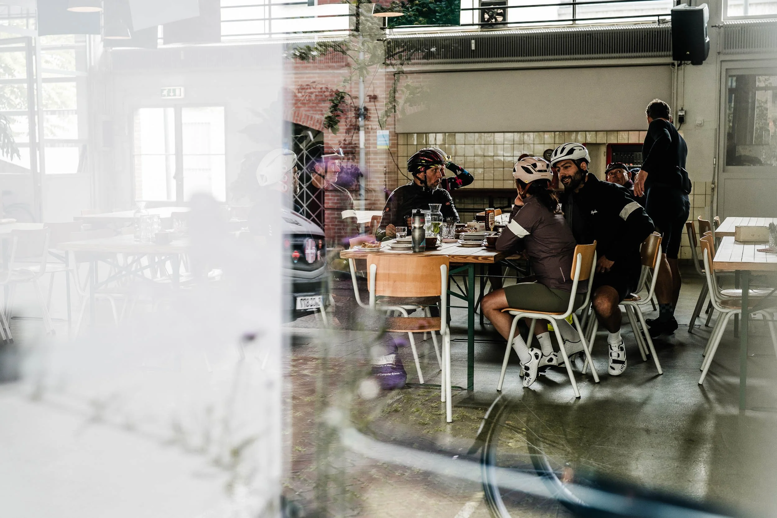 Group of cyclists wearing helmets and sportswear sitting at a table inside a cafe, seen through a glass window.