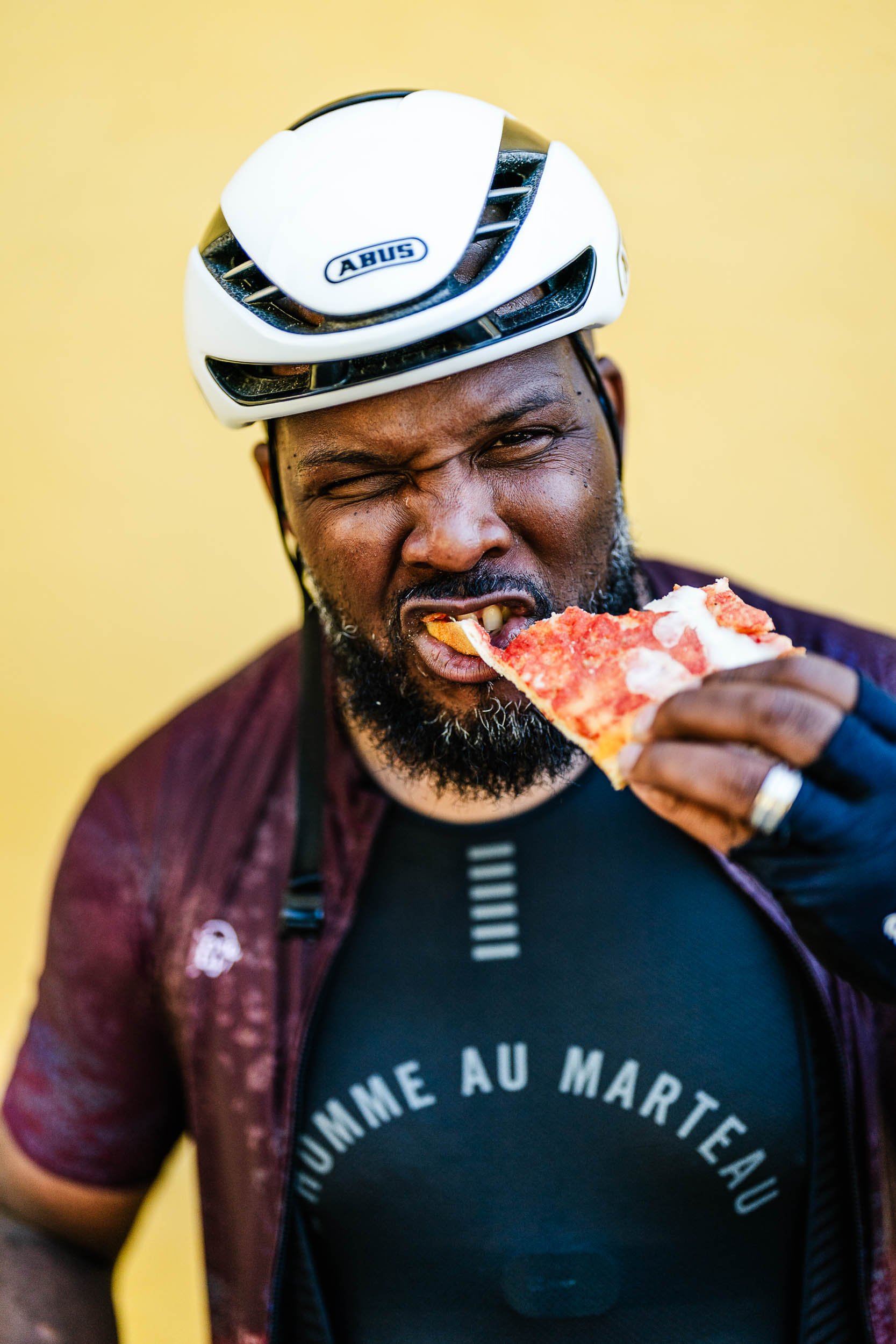 A man wearing a bicycle helmet and cycling gear eats a slice of pizza.